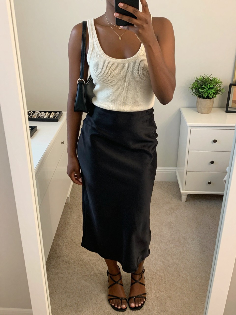 mirror selfie of a black woman wearing a cream knit tank top with a black satin slip skirt, black strappy flat sandals, a small black shoulder bag, and delicate layered gold necklaces, clearly showing the same clothing items, colors, fabrics, and accessories listed in the Outfit Pieces section, holding a phone in front of a mirror, cropped at NECK so the face does not show, with a relaxed casual selfie pose, in a feminine-themed bedroom, soft indoor lighting, including a jewelry tray on the vanity and a small plant on a dresser, latest iPhone photo quality.