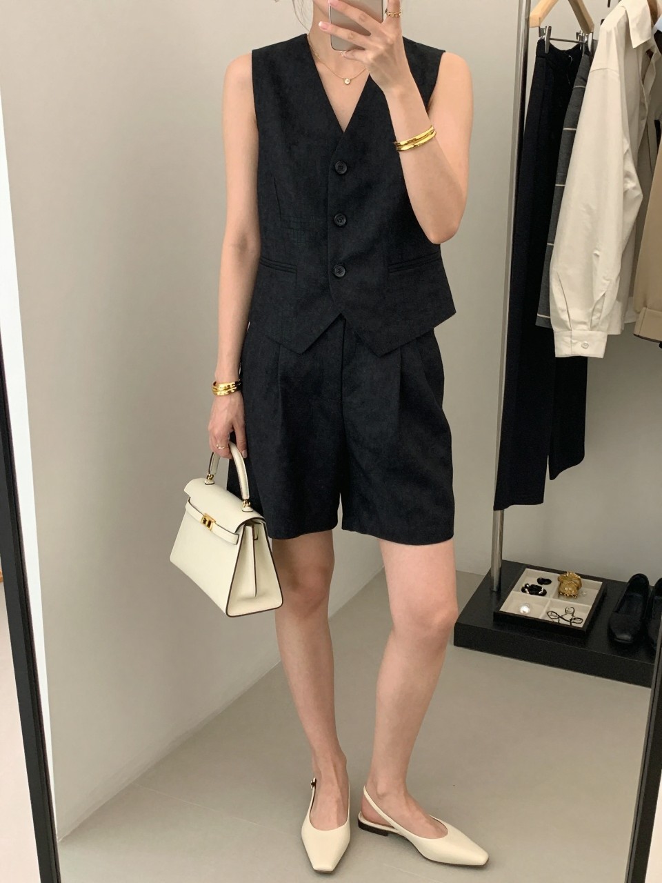 mirror selfie of a woman wearing a black linen waistcoat vest with matching black tailored linen shorts, ivory slingback flats, a small ivory top-handle bag, and gold cuff bracelets, clearly showing the same clothing items, colors, fabrics, and accessories listed in the Outfit Pieces section, holding a phone in front of a mirror, cropped at NECK so the face does not show, with a relaxed casual selfie pose, in a fashion studio mirror corner, soft indoor lighting, including a clothing rack with garments behind and a jewelry tray on the vanity, latest iPhone photo quality.