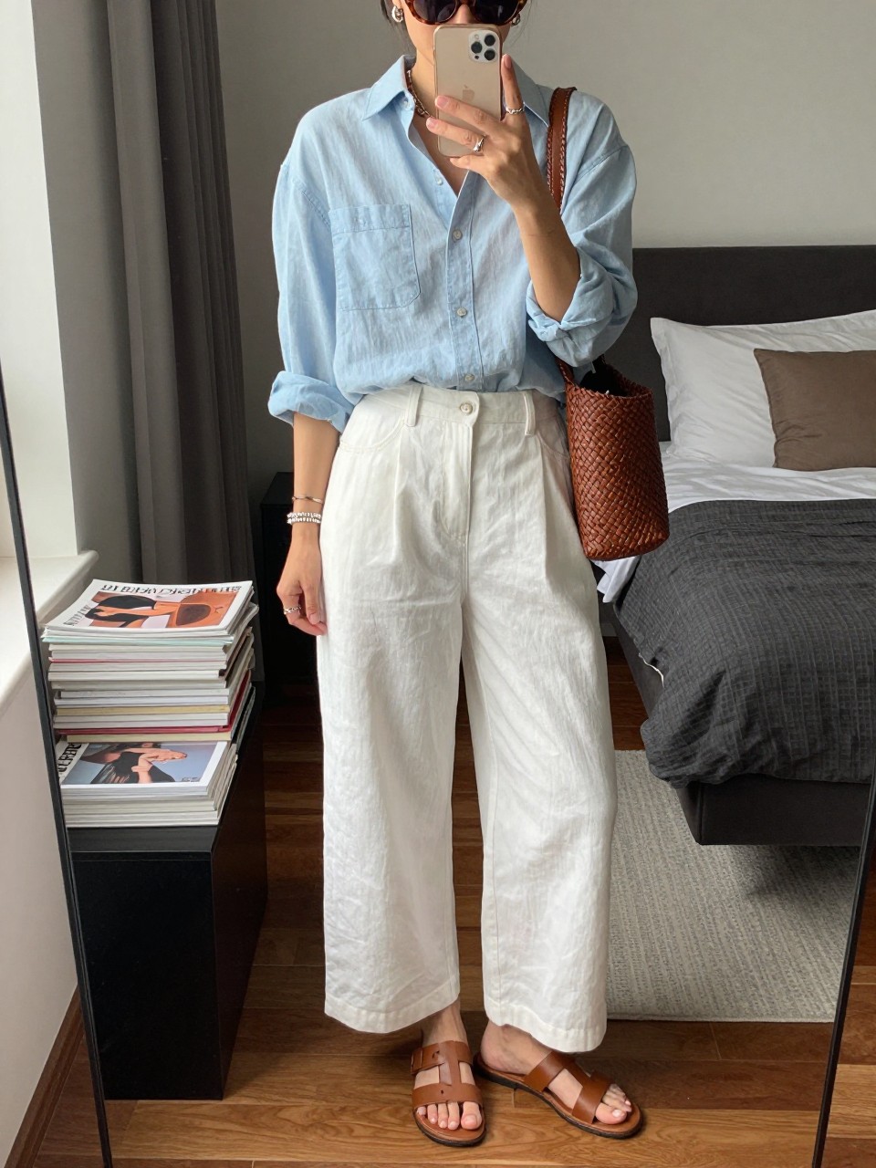 IMAGE_PROMPT: mirror selfie of a woman wearing a light chambray button-down shirt, white linen wide-leg pants, tan leather flat sandals, brown woven crossbody bag, and tortoiseshell sunglasses, clearly showing the same clothing items listed in the Outfit Pieces section, holding a phone in front of a mirror, cropped at the NECK so the face does not show, relaxed casual selfie pose, in a stylish city apartment bedroom, natural window light, with stack of fashion magazines on a side table, latest iPhone photo quality.