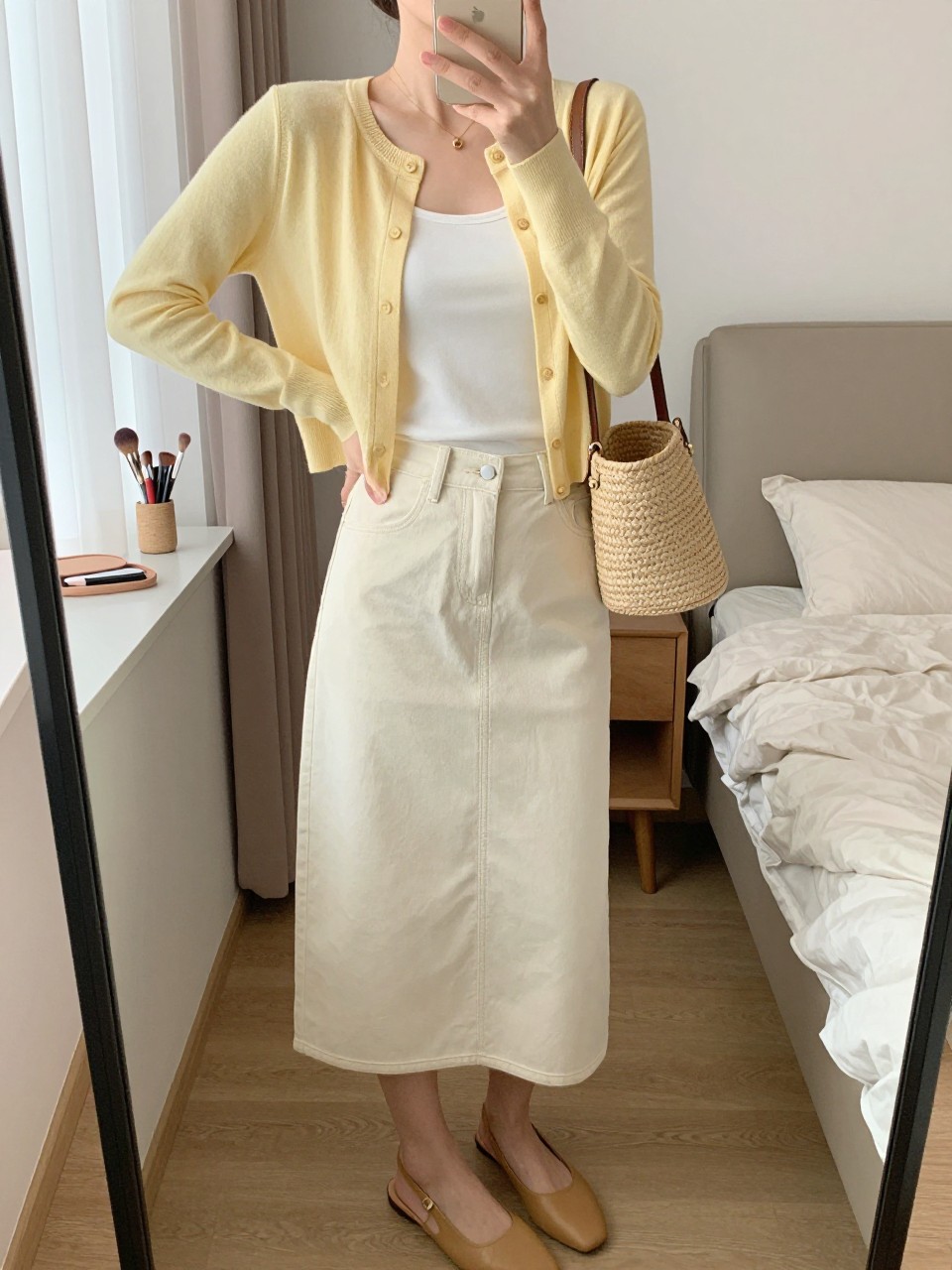 mirror selfie of a woman wearing a butter yellow cropped cardigan buttoned over a white fitted tank, cream denim midi skirt, tan slingback flats, a straw mini bag, and tiny gold studs, clearly showing the same clothing items, colors, fabrics, and accessories listed in the Outfit Pieces section, holding a phone in front of a mirror, cropped at NECK so the face does not show, with a relaxed casual selfie pose, in a feminine-themed bedroom, natural window light, including a makeup brush or cosmetic bag on the counter and a folded blanket on a chair, latest iPhone photo quality.