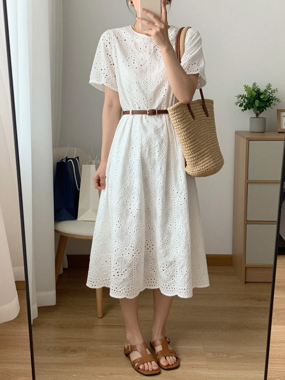 mirror selfie of a woman wearing a white eyelet midi dress, tan leather flat sandals, a natural raffia tote bag, a slim brown belt, and small gold hoop earrings, clearly showing the same clothing items, colors, fabrics, and accessories listed in the Outfit Pieces section, holding a phone in front of a mirror, cropped at NECK so the face does not show, with a relaxed casual selfie pose, in a cozy bedroom mirror setup, natural window light, including shopping bags beside a chair and a small plant on a dresser, latest iPhone photo quality.