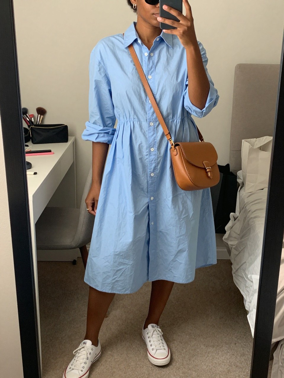 mirror selfie of a black woman wearing a light blue poplin shirt dress with white low-top sneakers, a tan crossbody saddle bag, black oval sunglasses, and small silver hoop earrings, clearly showing the same clothing items, colors, fabrics, and accessories listed in the Outfit Pieces section, holding a phone in front of a mirror, cropped at NECK so the face does not show, with a relaxed casual selfie pose, in a cozy bedroom mirror setup, soft indoor lighting, including a makeup brush or cosmetic bag on the counter and shopping bags beside a chair, latest iPhone photo quality.