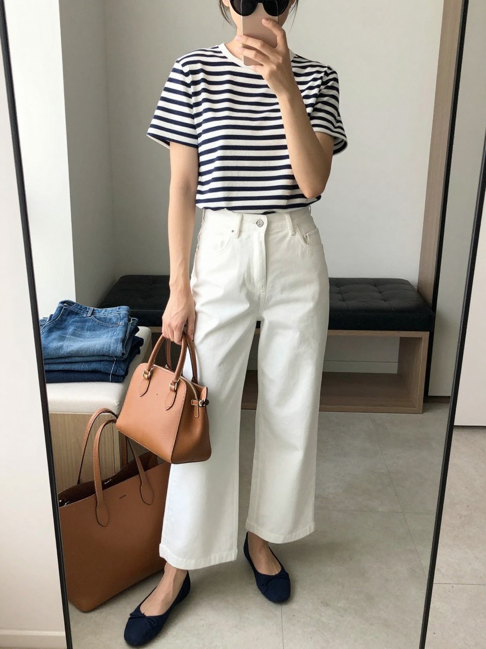 mirror selfie of a woman wearing a navy and white striped cotton tee tucked into white wide-leg trousers, navy ballet flats, a structured tan mini tote, and black cat-eye sunglasses, clearly showing the same clothing items, colors, fabrics, and accessories listed in the Outfit Pieces section, holding a phone in front of a mirror, cropped at NECK so the face does not show, with a relaxed casual selfie pose, in a walk-in closet mirror, natural window light, including a tote bag resting on the floor and folded denim jeans on a bench, latest iPhone photo quality.