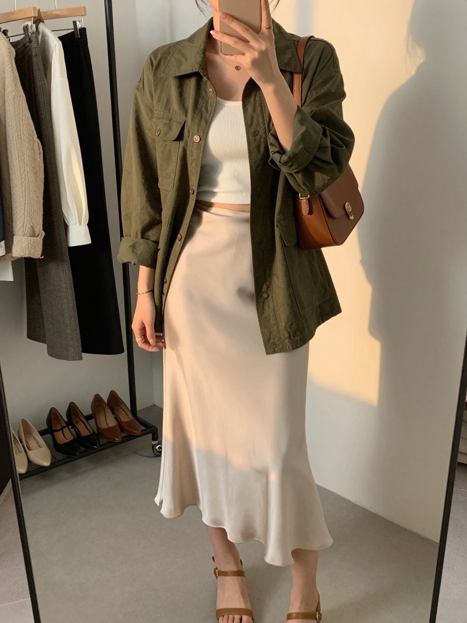 IMAGE_PROMPT: mirror selfie of a woman wearing an olive green lightweight utility jacket, white ribbed fitted tank, cream satin midi skirt, tan ankle-strap sandals, and brown mini shoulder bag, holding a phone in front of a mirror, cropped at the NECK so the face does not show, relaxed casual selfie pose, in a fashion studio mirror corner, evening golden hour, with clothing rack behind and heels on the floor nearby. Latest iPhone photo quality.
