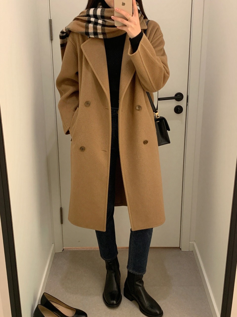 mirror selfie of a woman wearing a camel wool-blend coat over a black ribbed turtleneck, dark wash slim straight jeans, black leather ankle boots, and a plaid scarf in camel, black, and cream tones, clearly showing the same clothing items, colors, fabrics, and accessories listed in the Outfit Pieces section, holding a phone in front of a mirror, cropped at NECK so the face does not show, with a relaxed casual selfie pose, in a minimal dressing room, evening golden hour, including a handbag hanging on a door handle and a pair of heels on the floor nearby, latest iPhone photo quality.
