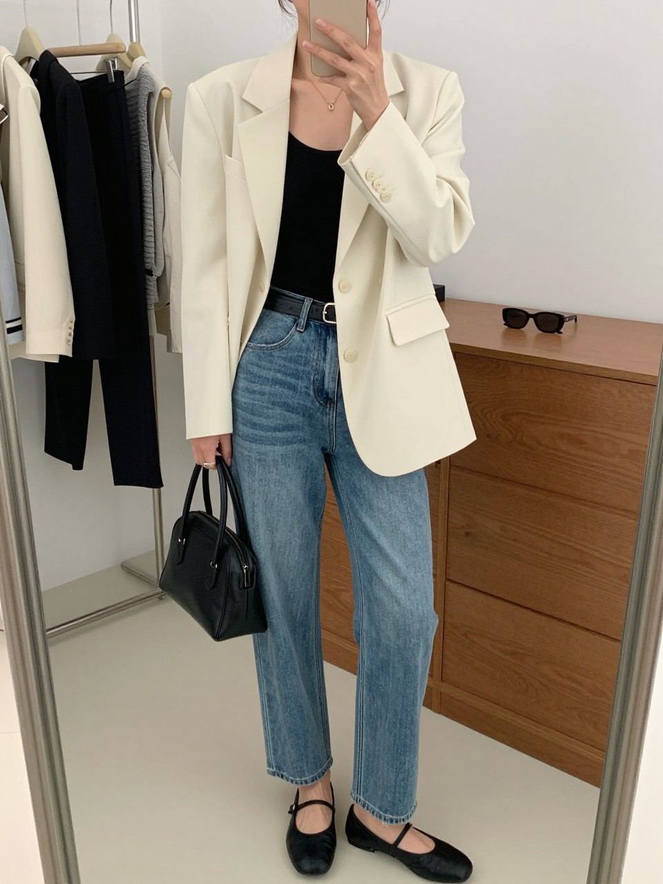 mirror selfie of a woman wearing a cream lightweight blazer over a black fitted tank, vintage blue straight-leg jeans, black ballet flats, a black leather belt, and a structured black mini bag, clearly showing the same clothing items, colors, fabrics, and accessories listed in the Outfit Pieces section, holding a phone in front of a mirror, cropped at NECK so the face does not show, with a relaxed casual selfie pose, in a fashion studio mirror corner, natural window light, including a clothing rack with garments behind and sunglasses placed on a dresser, latest iPhone photo quality.