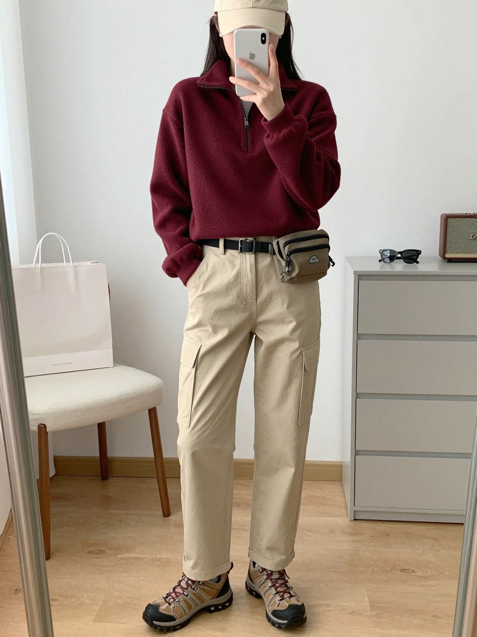 mirror selfie of a woman wearing a burgundy fleece pullover with beige straight-leg cargo pants, brown waterproof hiking shoes, a cream baseball cap, and a small nylon belt bag, clearly showing the same clothing items, colors, fabrics, and accessories listed in the Outfit Pieces section, holding a phone in front of a mirror, cropped at NECK so the face does not show, with a relaxed casual selfie pose, in a minimal dressing room, natural window light, including shopping bags beside a chair and sunglasses placed on a dresser, latest iPhone photo quality.