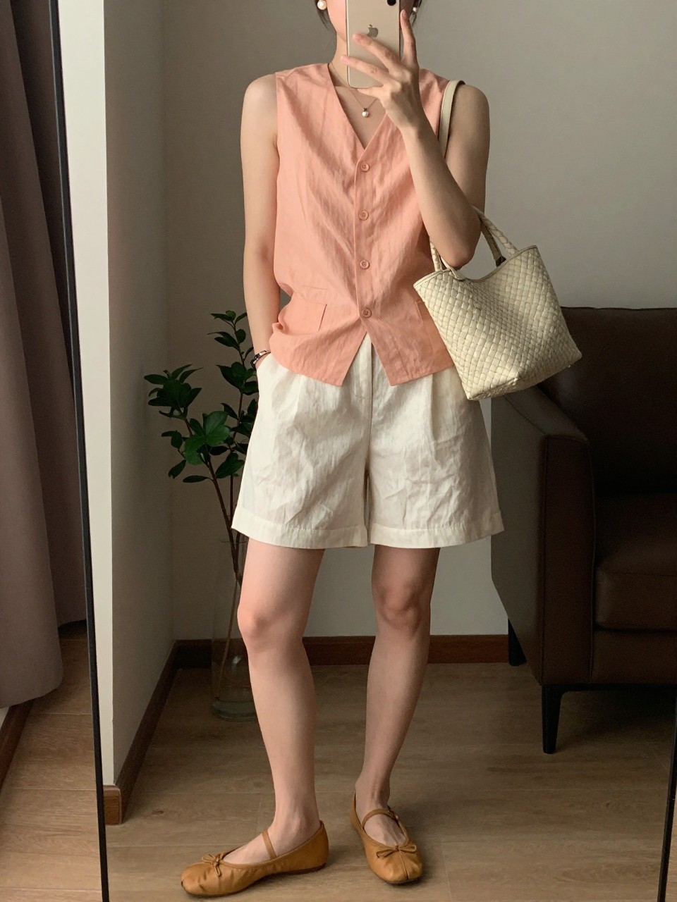 IMAGE_PROMPT: mirror selfie of a woman wearing a peach button-front sleeveless top, cream linen tailored shorts, tan leather ballet flats, cream woven handbag, and pearl stud earrings, clearly showing the same clothing items listed in the Outfit Pieces section, holding a phone in front of a mirror, cropped at the NECK so the face does not show, relaxed casual selfie pose, in a modern living room mirror setup, warm moody lighting, with small plant beside the floor mirror, latest iPhone photo quality.