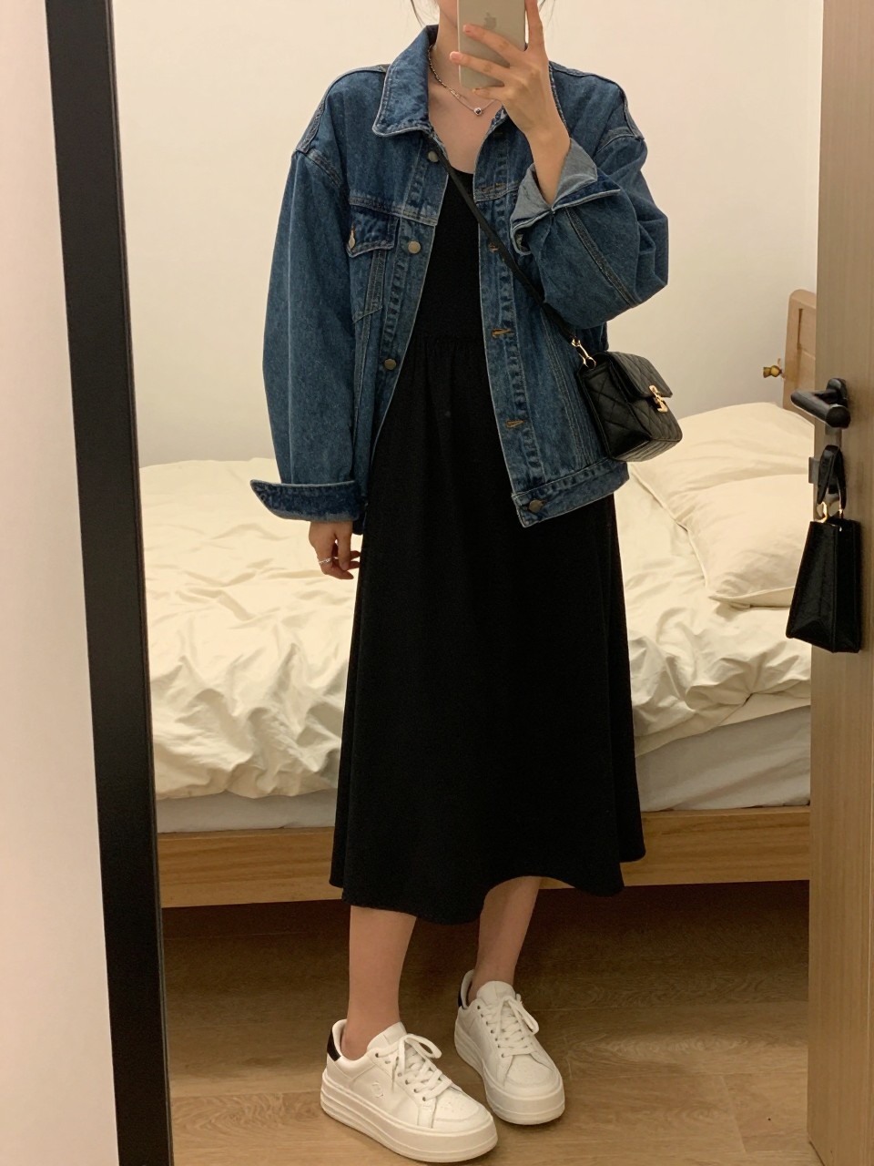 IMAGE_PROMPT: mirror selfie of a woman wearing a sleeveless black cotton midi dress, oversized blue denim jacket, white platform sneakers, black quilted crossbody bag, and small silver hoops, holding a phone in front of a mirror, cropped at the NECK so the face does not show, relaxed casual selfie pose, in a cozy bedroom mirror setup, warm moody lighting, with soft pillows on an unmade bed and handbag on a door handle. Latest iPhone photo quality.