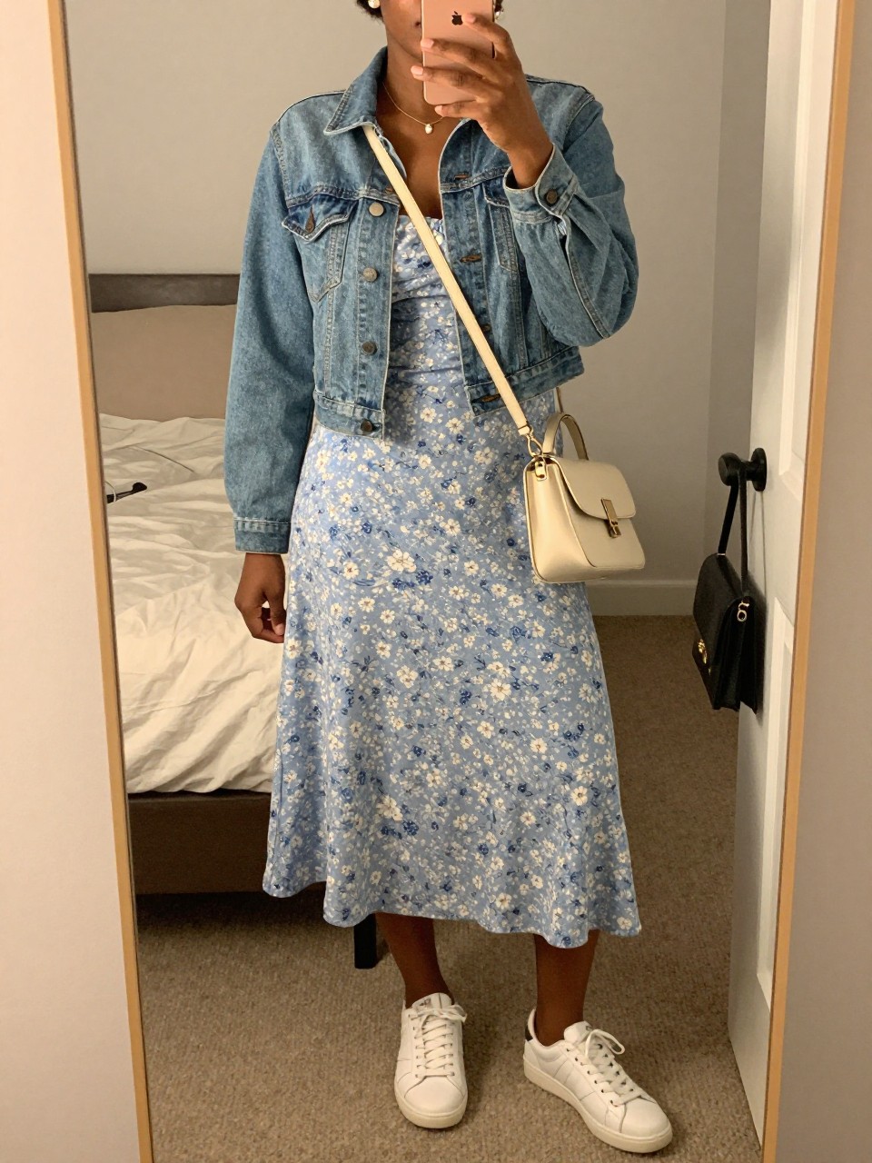 mirror selfie of a black woman wearing a soft blue floral midi dress with a cropped light wash denim jacket, white leather sneakers, a cream crossbody bag, and small pearl earrings, clearly showing the same clothing items, colors, fabrics, and accessories listed in the Outfit Pieces section, holding a phone in front of a mirror, cropped at NECK so the face does not show, with a relaxed casual selfie pose, in a cozy bedroom mirror setup, evening golden hour, including soft pillows on an unmade bed and a handbag hanging on a door handle, latest iPhone photo quality.