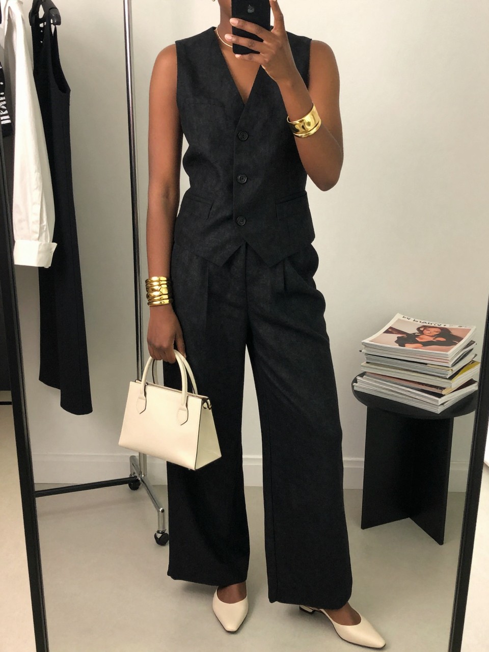 mirror selfie of a black woman wearing a black linen waistcoat vest with matching black linen wide-leg pants, ivory slingback flats, a small ivory top-handle bag, and gold cuff bracelets, clearly showing the same clothing items, colors, fabrics, and accessories listed in the Outfit Pieces section, holding a phone in front of a mirror, cropped at NECK so the face does not show, with a relaxed casual selfie pose, in a fashion studio mirror corner, soft indoor lighting, including a clothing rack with garments behind and a stack of fashion magazines on a side table, latest iPhone photo quality.