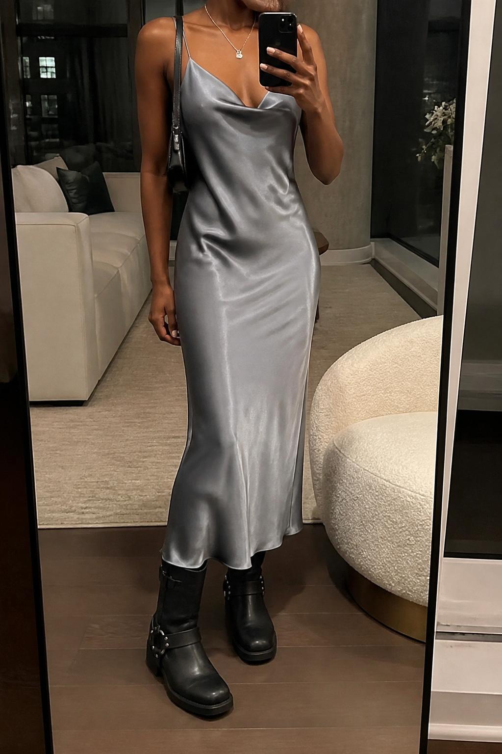IMAGE_PROMPT: mirror selfie of a black woman wearing a grey satin slip dress, black moto boots, small black shoulder bag, silver pendant necklace, and sleek low bun, clearly showing the same clothing items listed in the Outfit Pieces section, holding a phone in front of a mirror, cropped at the NECK so the face does not show, relaxed casual selfie pose, in a stylish city apartment living room, warm moody lighting, with accent chair beside the mirror, latest iPhone photo quality.