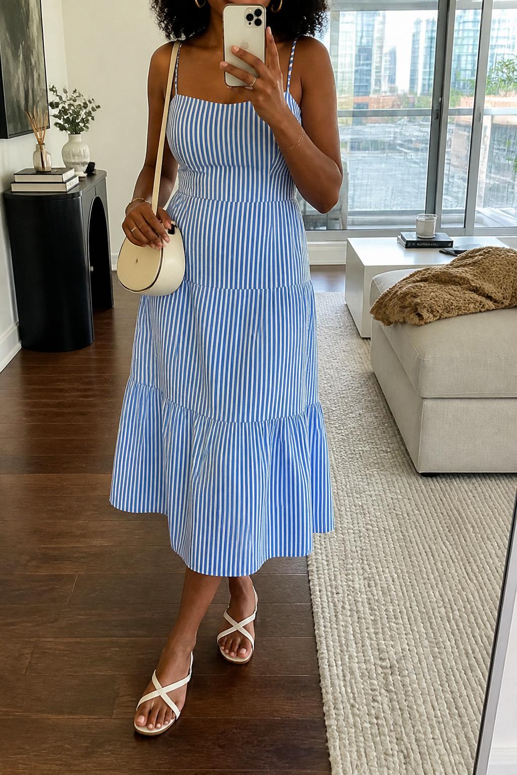 IMAGE_PROMPT: mirror selfie of a black woman wearing a blue and white striped cotton midi dress, white flat sandals, cream mini shoulder bag, small gold hoop earrings, and soft natural curls, clearly showing the same clothing items listed in the Outfit Pieces section, holding a phone in front of a mirror, cropped at the NECK so the face does not show, relaxed casual selfie pose, in a stylish city apartment living room, soft morning light, with throw blanket on a living room sofa, latest iPhone photo quality.