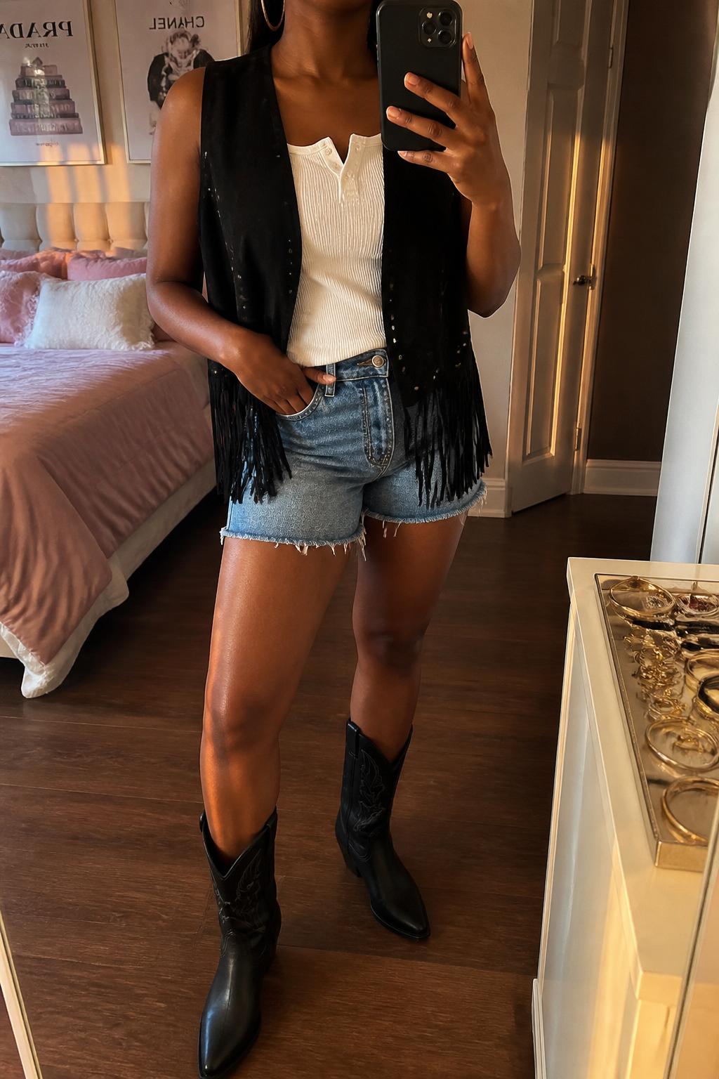 IMAGE_PROMPT: mirror selfie of a black woman wearing a black fringe suede vest, white ribbed tank top, cutoff medium wash denim shorts, black western boots, and silver hoop earrings, clearly showing the same clothing items listed in the Outfit Pieces section, holding a phone in front of a mirror, cropped at the NECK so the face does not show, relaxed casual selfie pose, in a feminine-themed bedroom, evening golden hour, with jewelry tray on the vanity, latest iPhone photo quality.