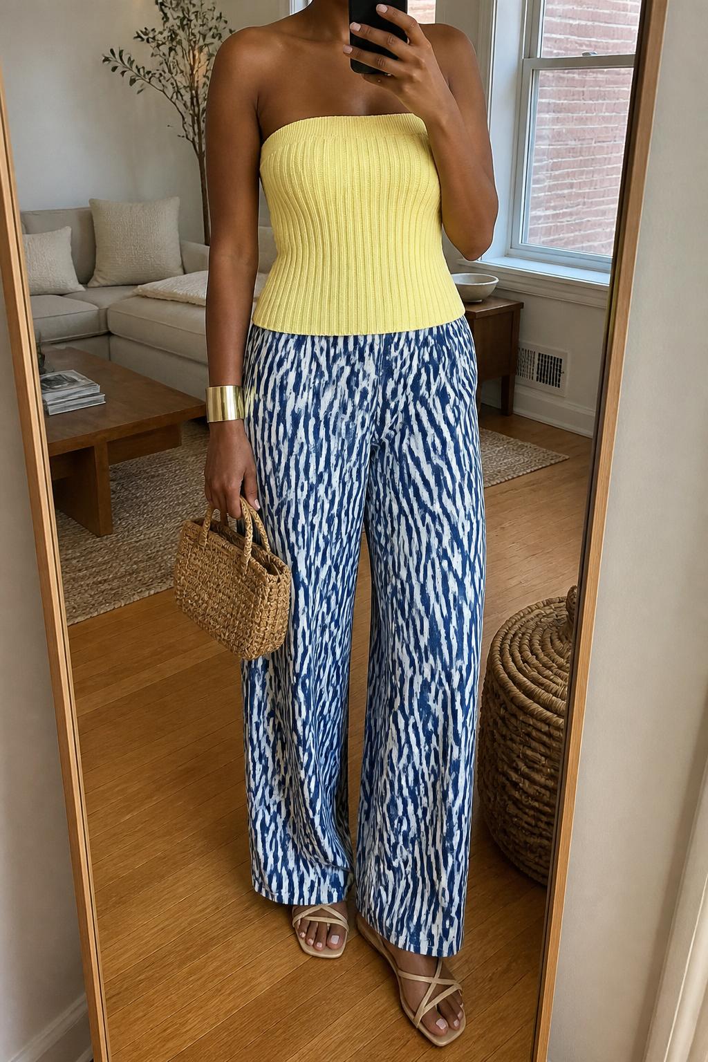 IMAGE_PROMPT: mirror selfie of a black woman wearing a butter yellow thick ribbed tube top, blue and white printed wide-leg pants, tan strappy flat sandals, woven mini handbag, and gold cuff bracelet, clearly showing the same clothing items listed in the Outfit Pieces section, holding a phone in front of a mirror, cropped at the NECK so the face does not show, relaxed casual selfie pose, in a cozy living room with a leaning floor mirror, natural window light, with woven basket near the mirror, latest iPhone photo quality.