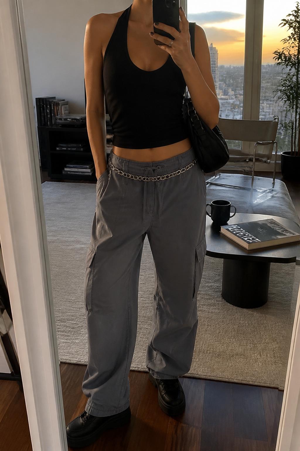 IMAGE_PROMPT: mirror selfie of a woman wearing a black cotton halter top, grey lightweight cargo trousers, black platform sneakers, silver chain belt, and black nylon shoulder bag, clearly showing the same clothing items listed in the Outfit Pieces section, holding a phone in front of a mirror, cropped at the NECK so the face does not show, relaxed casual selfie pose, in a stylish city apartment living room, evening golden hour, with coffee table with a book and mug, latest iPhone photo quality.