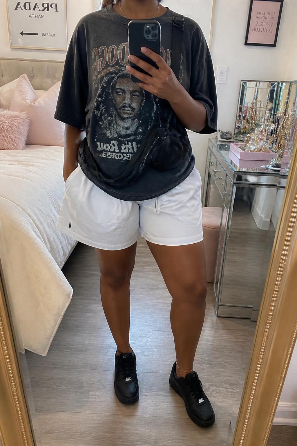 IMAGE_PROMPT: mirror selfie of a black woman wearing an oversized faded charcoal graphic tee, white cotton boxer shorts, black low-top sneakers, black belt bag, and messy low bun, clearly showing the same clothing items listed in the Outfit Pieces section, holding a phone in front of a mirror, cropped at the NECK so the face does not show, relaxed casual selfie pose, in a feminine-themed bedroom, soft indoor lighting, with jewelry tray on the vanity, latest iPhone photo quality.