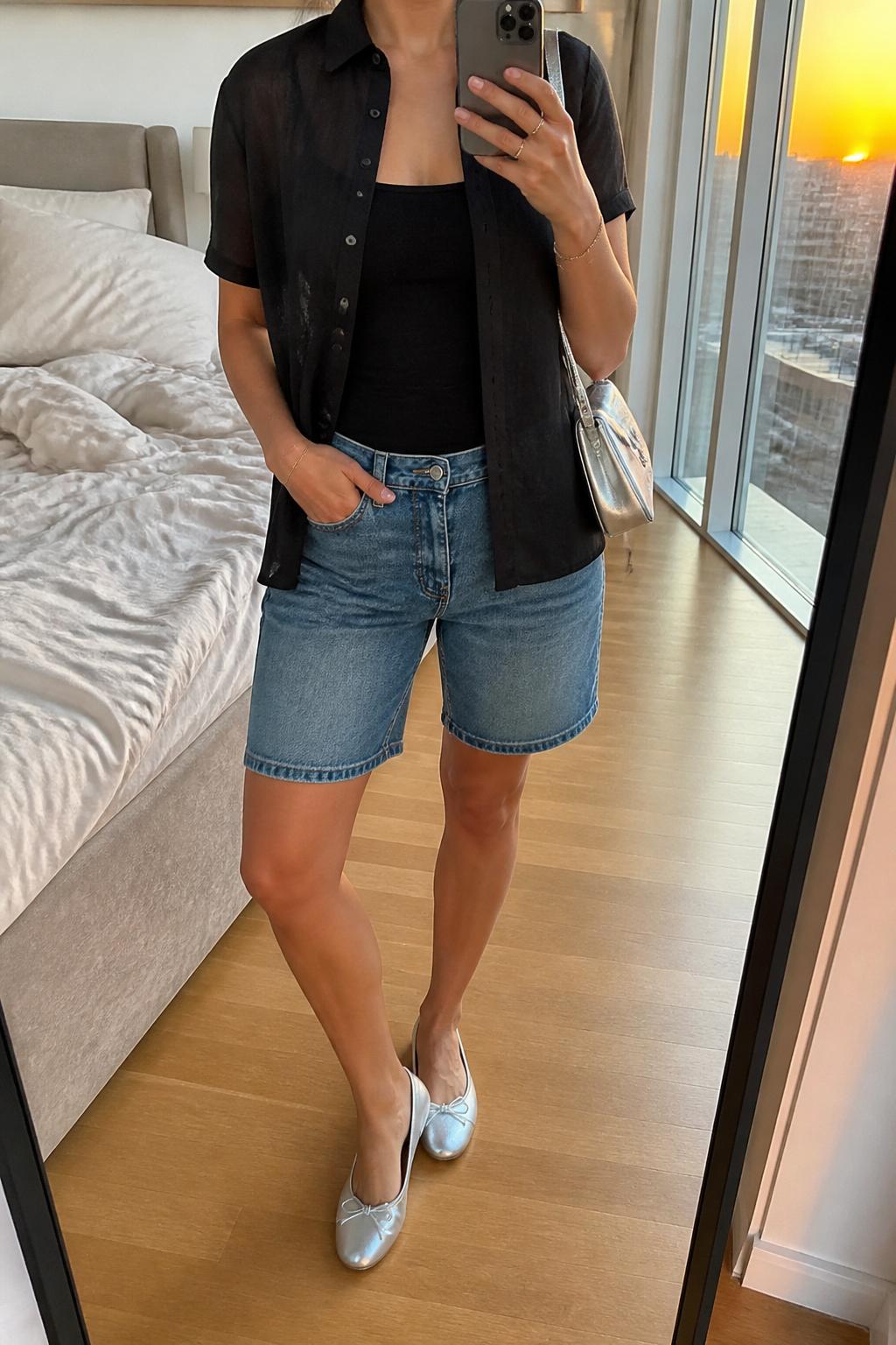 IMAGE_PROMPT: mirror selfie of a woman wearing a sheer black short-sleeve button-down shirt, black fitted tank top, medium wash denim Bermuda shorts, silver ballet flats, and small silver shoulder bag, clearly showing the same clothing items listed in the Outfit Pieces section, holding a phone in front of a mirror, cropped at the NECK so the face does not show, relaxed casual selfie pose, in a stylish city apartment bedroom, evening golden hour, with soft pillows on an unmade bed, latest iPhone photo quality.