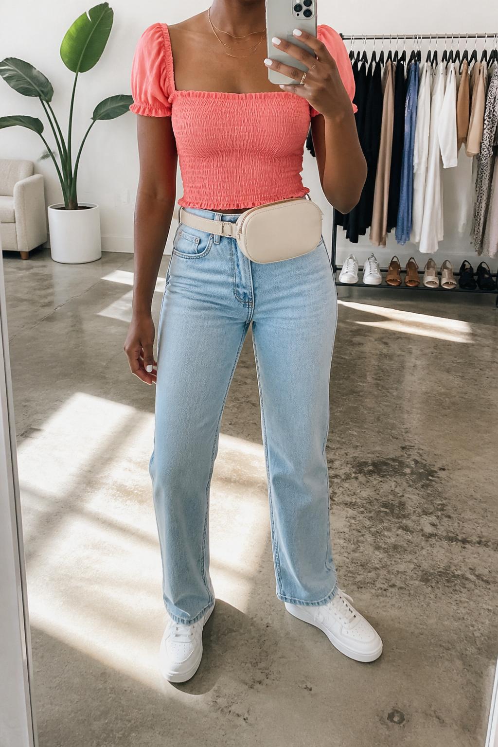 IMAGE_PROMPT: mirror selfie of a black woman wearing a coral smocked square-neck top, light wash straight-leg jeans, white leather sneakers, cream belt bag, and glossy pink lip makeup, clearly showing the same clothing items listed in the Outfit Pieces section, holding a phone in front of a mirror, cropped at the NECK so the face does not show, relaxed casual selfie pose, in a fashion studio mirror corner, soft morning light, with clothing rack with garments behind, latest iPhone photo quality.