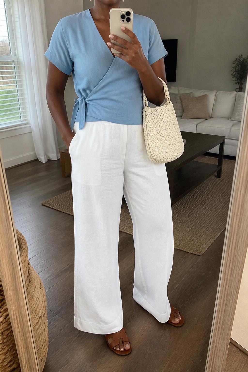 IMAGE_PROMPT: mirror selfie of a black woman wearing a soft blue short-sleeve wrap top, white wide-leg linen pants, tan leather flat sandals, cream woven handbag, and small pearl earrings, clearly showing the same clothing items listed in the Outfit Pieces section, holding a phone in front of a mirror, cropped at the NECK so the face does not show, relaxed casual selfie pose, in a cozy living room with a leaning floor mirror, natural window light, with woven basket near the mirror, latest iPhone photo quality.