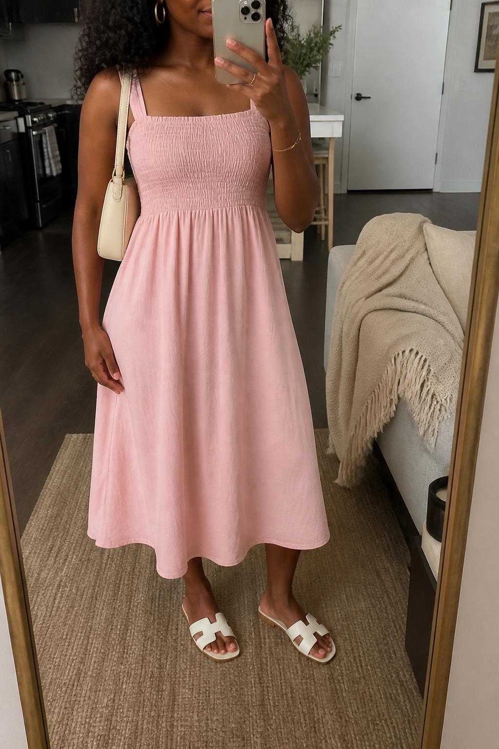 IMAGE_PROMPT: mirror selfie of a black woman wearing a soft pink smocked cotton midi dress, white flat sandals, cream mini shoulder bag, small gold hoop earrings, and soft natural curls, clearly showing the same clothing items listed in the Outfit Pieces section, holding a phone in front of a mirror, cropped at the NECK so the face does not show, relaxed casual selfie pose, in a stylish city apartment living room, soft morning light, with throw blanket on a living room sofa, latest iPhone photo quality.