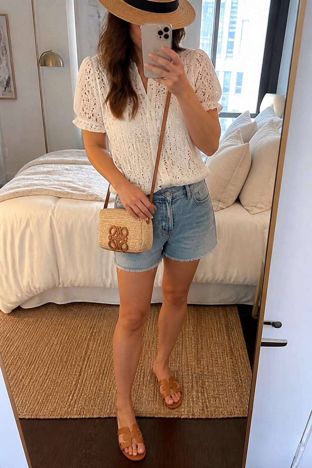 IMAGE_PROMPT: mirror selfie of a woman wearing a tan straw fedora hat, white eyelet short-sleeve blouse, light wash denim shorts, brown leather sandals, and raffia crossbody bag, clearly showing the same clothing items listed in the Outfit Pieces section, holding a phone in front of a mirror, cropped at the NECK so the face does not show, relaxed casual selfie pose, in a stylish city apartment bedroom, soft indoor lighting, with soft pillows on an unmade bed, latest iPhone photo quality.