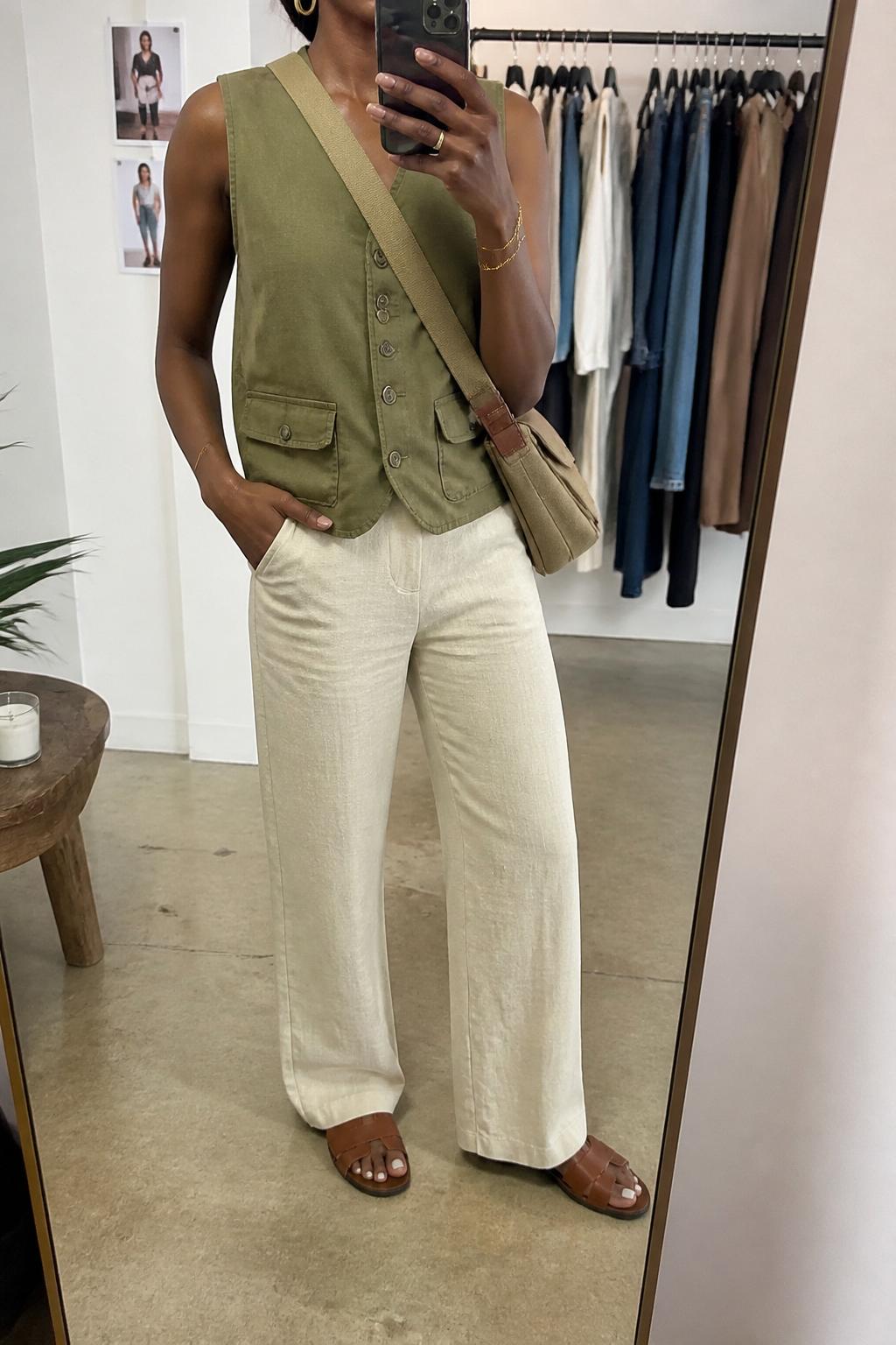 IMAGE_PROMPT: mirror selfie of a black woman wearing an olive utility vest, cream linen straight-leg pants, brown leather slide sandals, tan canvas crossbody bag, and small gold hoops, clearly showing the same clothing items listed in the Outfit Pieces section, holding a phone in front of a mirror, cropped at the NECK so the face does not show, relaxed casual selfie pose, in a fashion studio mirror corner, soft indoor lighting, with clothing rack with garments behind, latest iPhone photo quality.