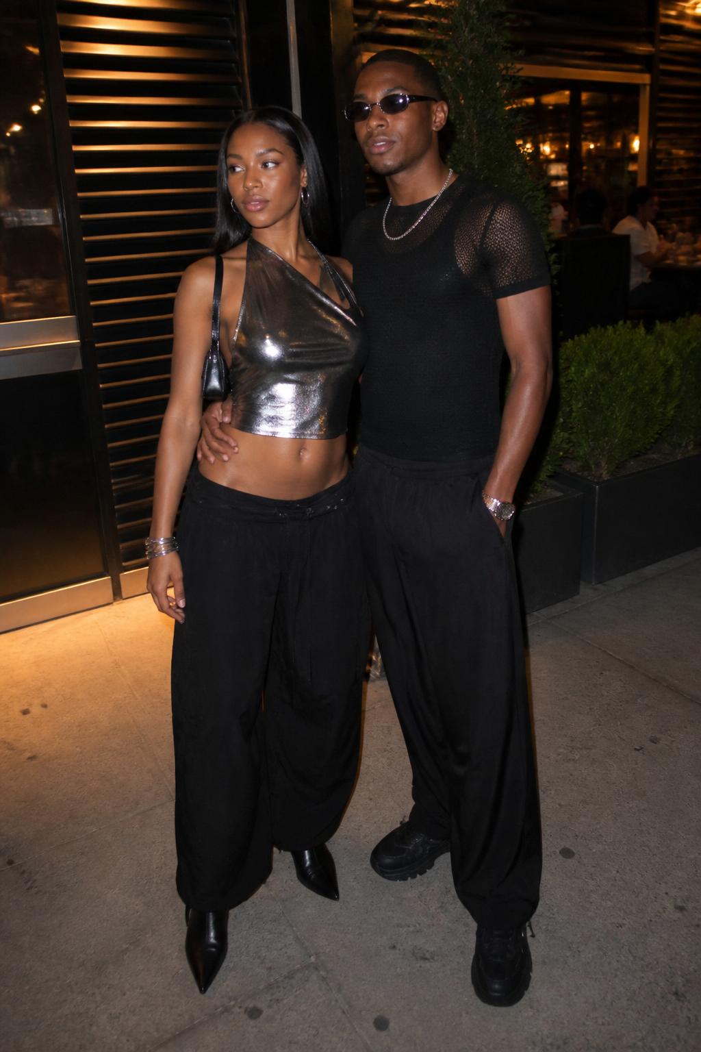Outfit Image Prompt:
A fashionable Black couple wearing Y2K night-out outfits near a casual downtown restaurant, woman wearing a silver metallic halter top, black low-rise baggy pants, black pointed boots, thin silver bangles, glossy but natural makeup, and a tiny black shoulder bag, man wearing a black fitted mesh shirt layered over a tank, black baggy trousers, silver chain necklace, black sneakers, and slim sunglasses, coordinated silver and black Y2K couple outfit, real nighttime street photo with soft restaurant lights, natural skin texture, realistic shadows, fabric wrinkles, slightly grainy low-light feel, no over-sharpening, no plastic skin, no fake glossy finish, no watermarks on images