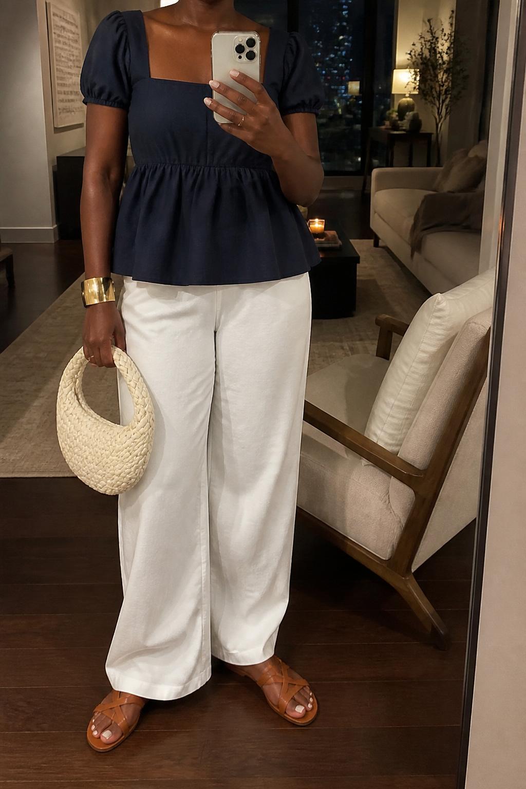 IMAGE_PROMPT: mirror selfie of a black woman wearing a navy cotton peplum top, white wide-leg pants, tan leather flat sandals, cream woven handbag, and gold cuff bracelet, clearly showing the same clothing items listed in the Outfit Pieces section, holding a phone in front of a mirror, cropped at the NECK so the face does not show, relaxed casual selfie pose, in a stylish city apartment living room, warm moody lighting, with accent chair beside the mirror, latest iPhone photo quality.