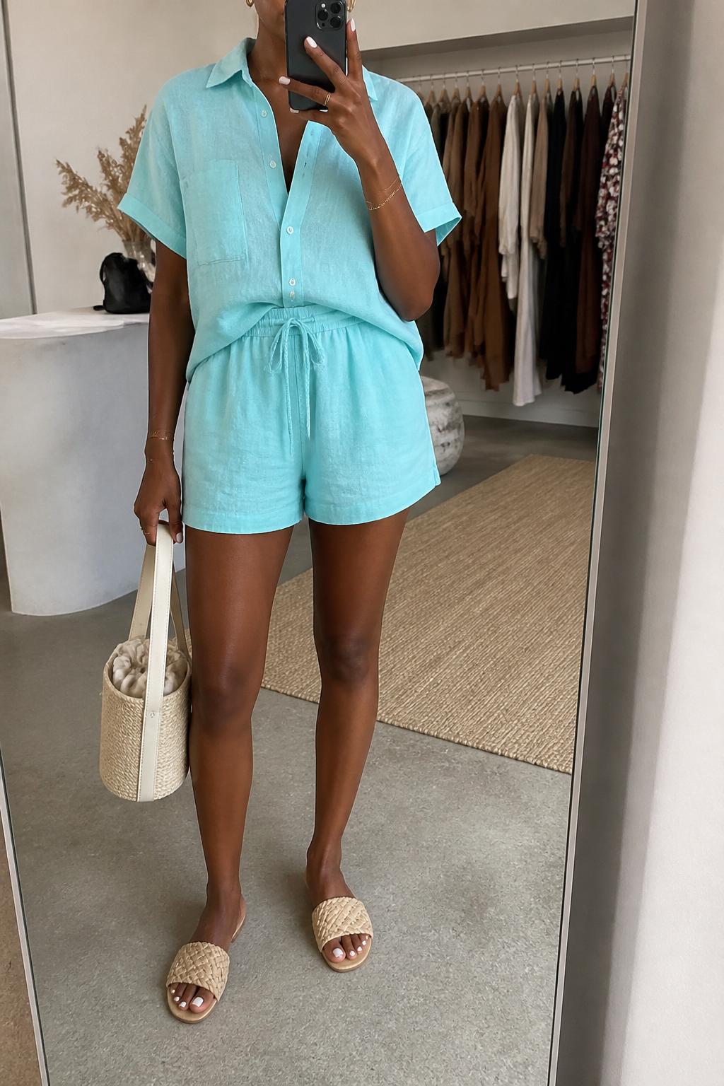 IMAGE_PROMPT: mirror selfie of a black woman wearing an aqua linen matching set with a short-sleeve button shirt and relaxed drawstring shorts, raffia slide sandals, cream bucket bag, and small gold hoops, clearly showing the same clothing items listed in the Outfit Pieces section, holding a phone in front of a mirror, cropped at the NECK so the face does not show, relaxed casual selfie pose, in a fashion studio mirror corner, soft indoor lighting, with clothing rack with garments behind, latest iPhone photo quality.