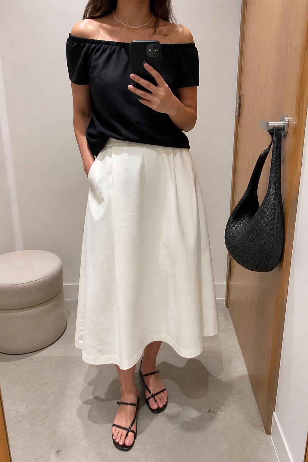 IMAGE_PROMPT: mirror selfie of a woman wearing a black off-shoulder cotton top, white cotton A-line midi skirt, black strappy flat sandals, woven black shoulder bag, and delicate pearl necklace, clearly showing the same clothing items listed in the Outfit Pieces section, holding a phone in front of a mirror, cropped at the NECK so the face does not show, relaxed casual selfie pose, in a minimal dressing room, soft indoor lighting, with handbag hanging on a door handle, latest iPhone photo quality.