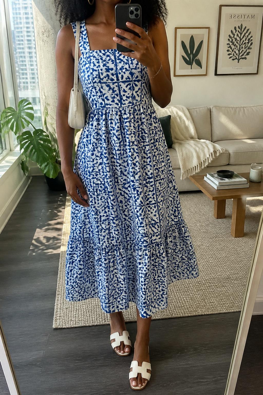 IMAGE_PROMPT: mirror selfie of a black woman wearing a blue tile-print cotton midi dress, white flat sandals, cream mini shoulder bag, small gold hoop earrings, and soft natural curls, clearly showing the same clothing items listed in the Outfit Pieces section, holding a phone in front of a mirror, cropped at the NECK so the face does not show, relaxed casual selfie pose, in a stylish city apartment living room, soft morning light, with throw blanket on a living room sofa, latest iPhone photo quality.
