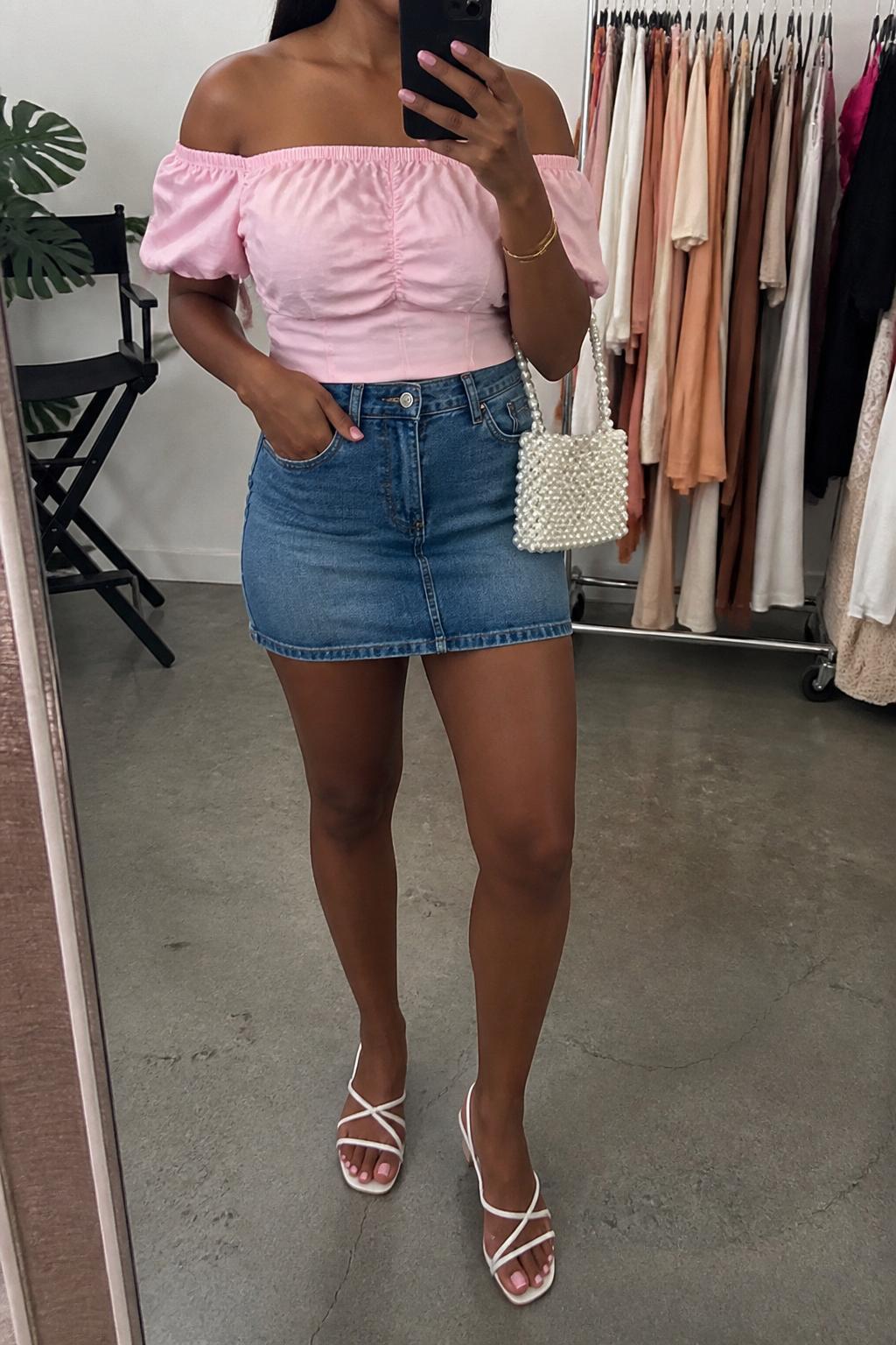 IMAGE_PROMPT: mirror selfie of a black woman wearing a soft pink off-shoulder cotton top, medium blue denim mini skirt, white strappy flat sandals, pearl mini bag, and glossy pink lip makeup, clearly showing the same clothing items listed in the Outfit Pieces section, holding a phone in front of a mirror, cropped at the NECK so the face does not show, relaxed casual selfie pose, in a fashion studio mirror corner, soft indoor lighting, with clothing rack with garments behind, latest iPhone photo quality.