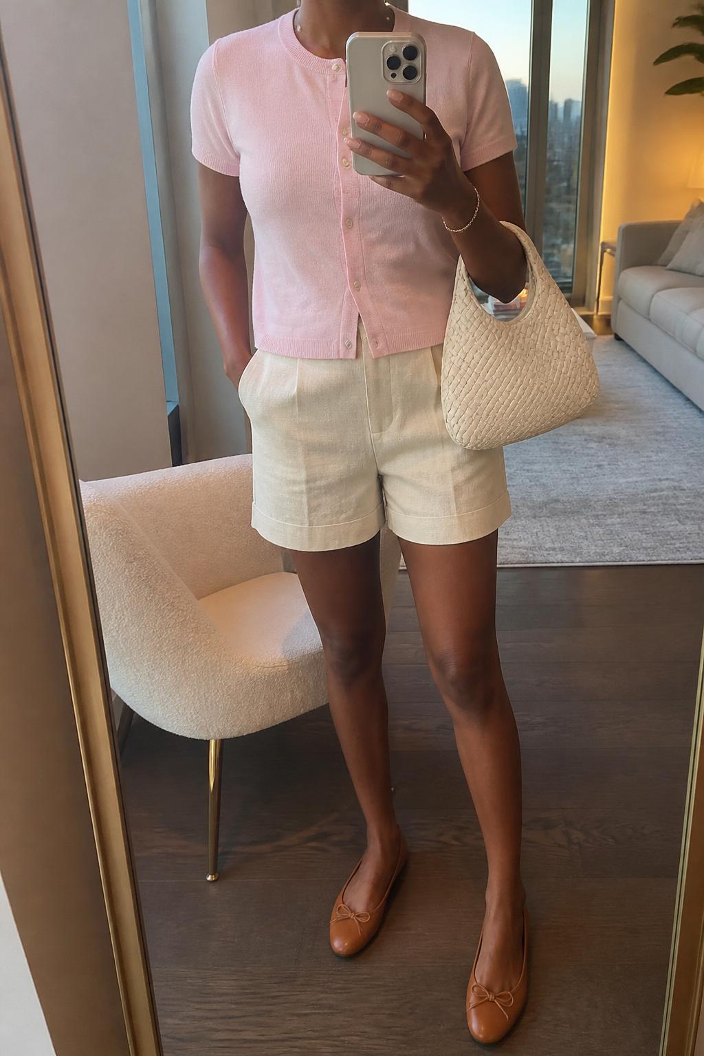 IMAGE_PROMPT: mirror selfie of a black woman wearing a pale pink lightweight short-sleeve cardigan, cream linen tailored shorts, tan leather ballet flats, cream woven handbag, and pearl stud earrings, clearly showing the same clothing items listed in the Outfit Pieces section, holding a phone in front of a mirror, cropped at the NECK so the face does not show, relaxed casual selfie pose, in a stylish city apartment living room, evening golden hour, with accent chair beside the mirror, latest iPhone photo quality.