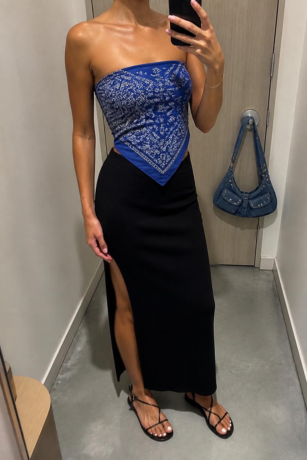 IMAGE_PROMPT: mirror selfie of a woman wearing a blue paisley bandana top, black cotton midi skirt with side slit, black strappy flat sandals, small denim shoulder bag, and glossy pink lip makeup, clearly showing the same clothing items listed in the Outfit Pieces section, holding a phone in front of a mirror, cropped at the NECK so the face does not show, relaxed casual selfie pose, in a minimal dressing room, soft indoor lighting, with handbag hanging on a door handle, latest iPhone photo quality.