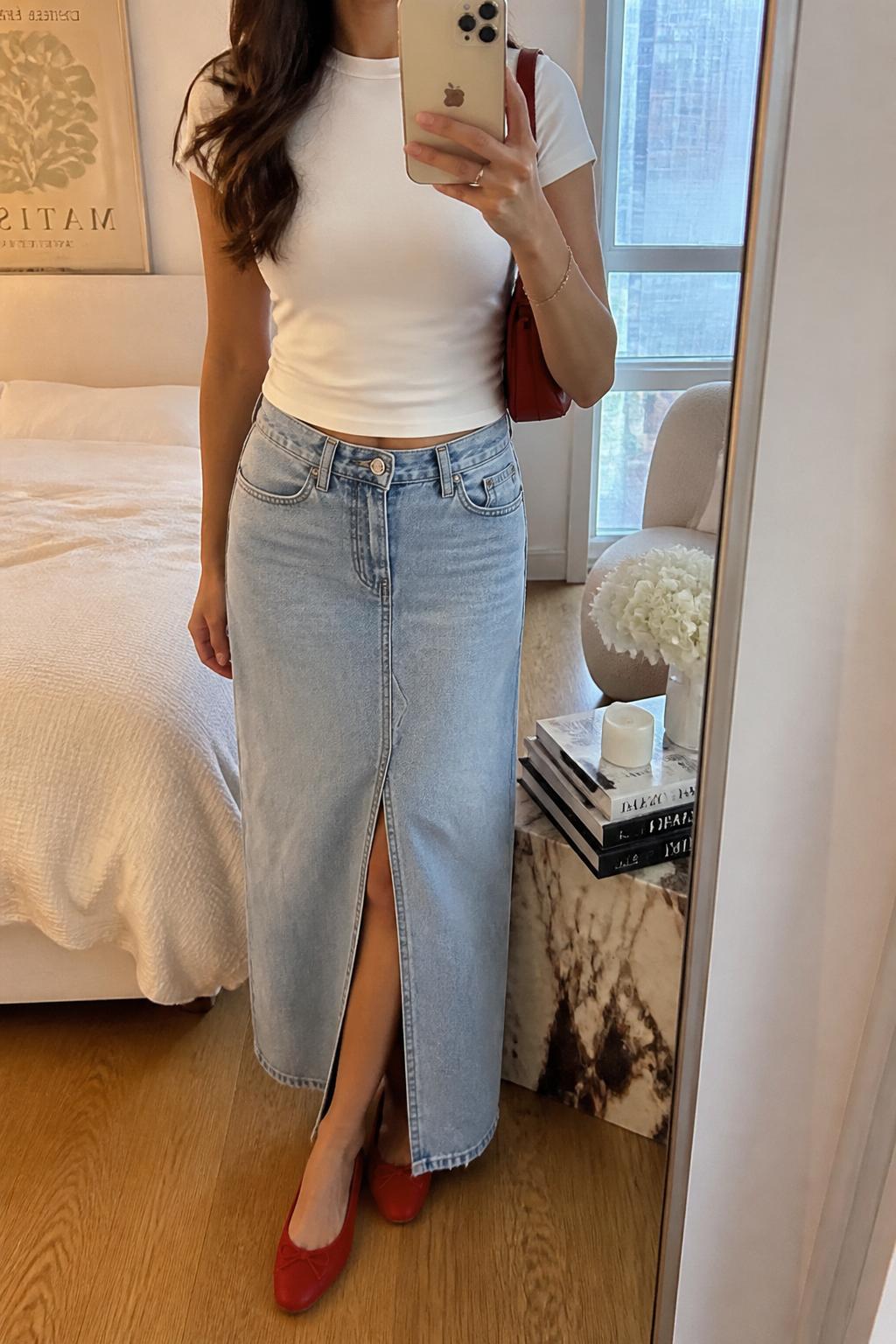 IMAGE_PROMPT: mirror selfie of a woman wearing a fitted white baby tee, light wash denim maxi skirt with front slit, red leather ballet flats, small red shoulder bag, and soft wavy hair, clearly showing the same clothing items listed in the Outfit Pieces section, holding a phone in front of a mirror, cropped at the NECK so the face does not show, relaxed casual selfie pose, in a stylish city apartment bedroom, soft indoor lighting, with stack of fashion magazines on a side table, latest iPhone photo quality.