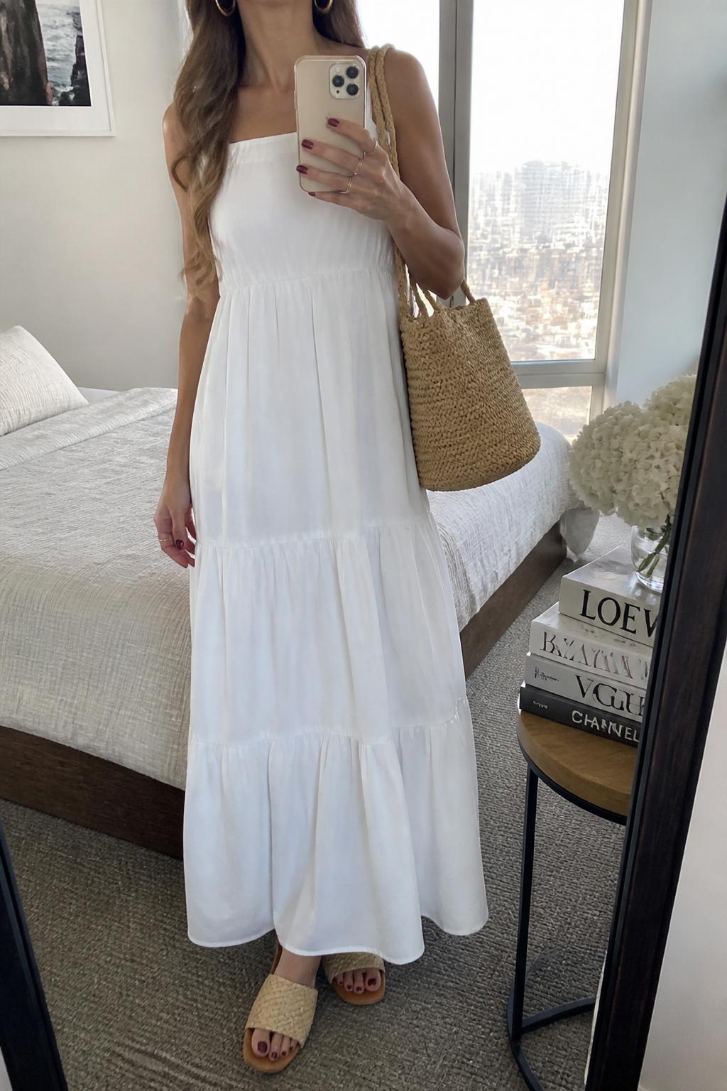 IMAGE_PROMPT: mirror selfie of a woman wearing a white cotton tiered maxi dress, raffia slide sandals, tan woven tote bag, small gold hoops, and loose wavy hair, clearly showing the same clothing items listed in the Outfit Pieces section, holding a phone in front of a mirror, cropped at the NECK so the face does not show, relaxed casual selfie pose, in a stylish city apartment bedroom, natural window light, with stack of fashion magazines on a side table, latest iPhone photo quality.