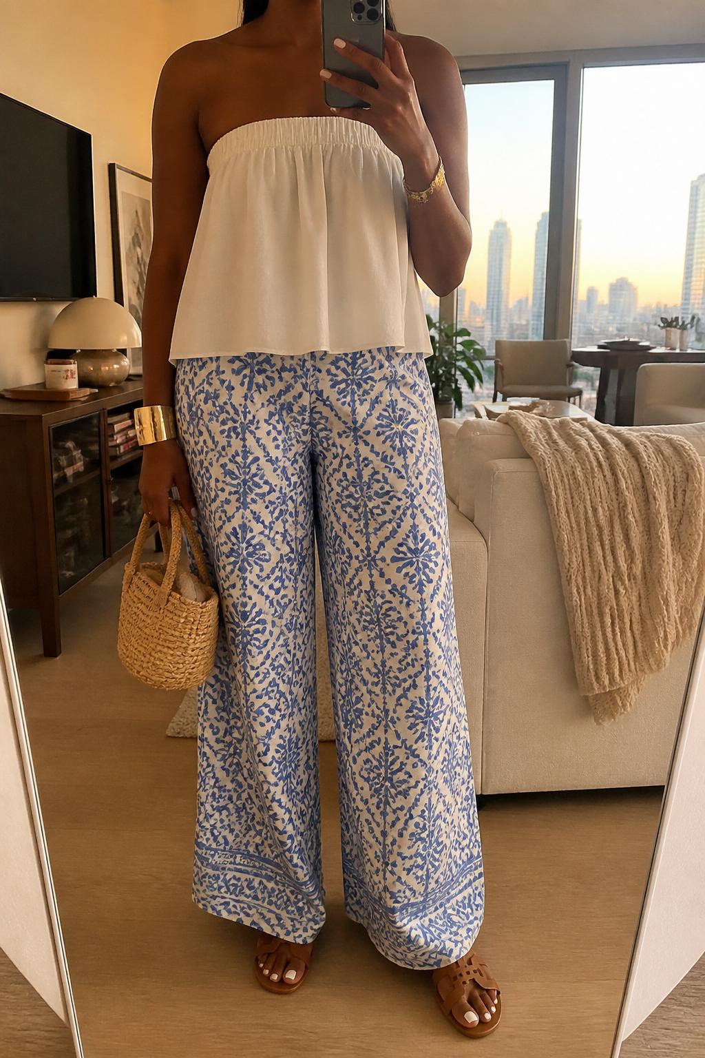 IMAGE_PROMPT: mirror selfie of a black woman wearing an ivory strapless cotton top, blue and white printed wide-leg pants, tan leather flat sandals, small raffia handbag, and gold cuff bracelet, clearly showing the same clothing items listed in the Outfit Pieces section, holding a phone in front of a mirror, cropped at the NECK so the face does not show, relaxed casual selfie pose, in a stylish city apartment living room, evening golden hour, with throw blanket on a living room sofa, latest iPhone photo quality.