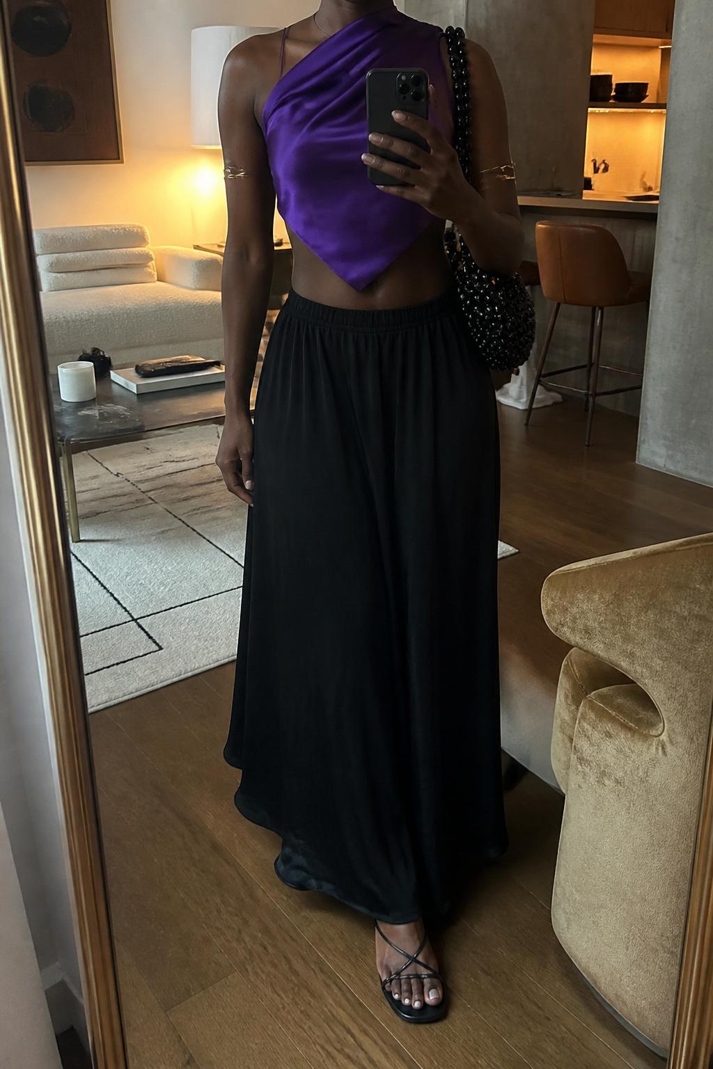 IMAGE_PROMPT: mirror selfie of a black woman wearing a purple satin scarf top, black flowy maxi skirt, black strappy flat sandals, beaded black shoulder bag, and gold arm cuff, clearly showing the same clothing items listed in the Outfit Pieces section, holding a phone in front of a mirror, cropped at the NECK so the face does not show, relaxed casual selfie pose, in a stylish city apartment living room, warm moody lighting, with accent chair beside the mirror, latest iPhone photo quality.