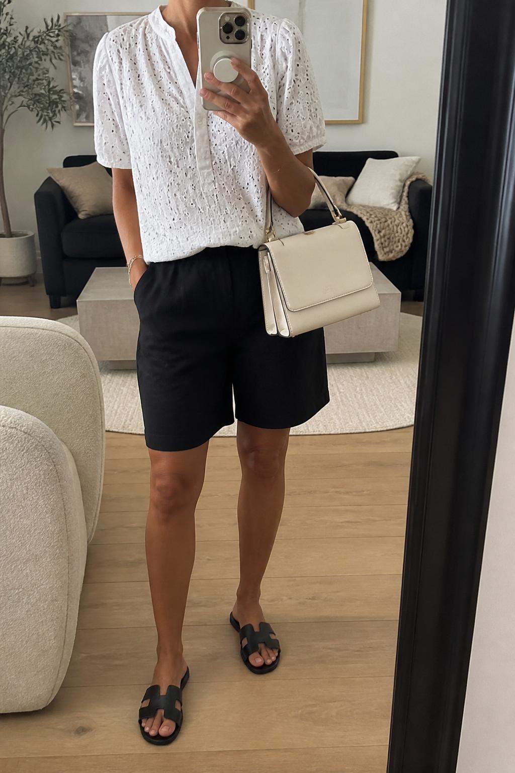 IMAGE_PROMPT: mirror selfie of a woman wearing a white eyelet short-sleeve blouse, black linen knee-length shorts, black leather flat sandals, cream top-handle bag, and glossy pink lip makeup, clearly showing the same clothing items listed in the Outfit Pieces section, holding a phone in front of a mirror, cropped at the NECK so the face does not show, relaxed casual selfie pose, in a modern living room mirror setup, soft indoor lighting, with accent chair beside the mirror, latest iPhone photo quality.