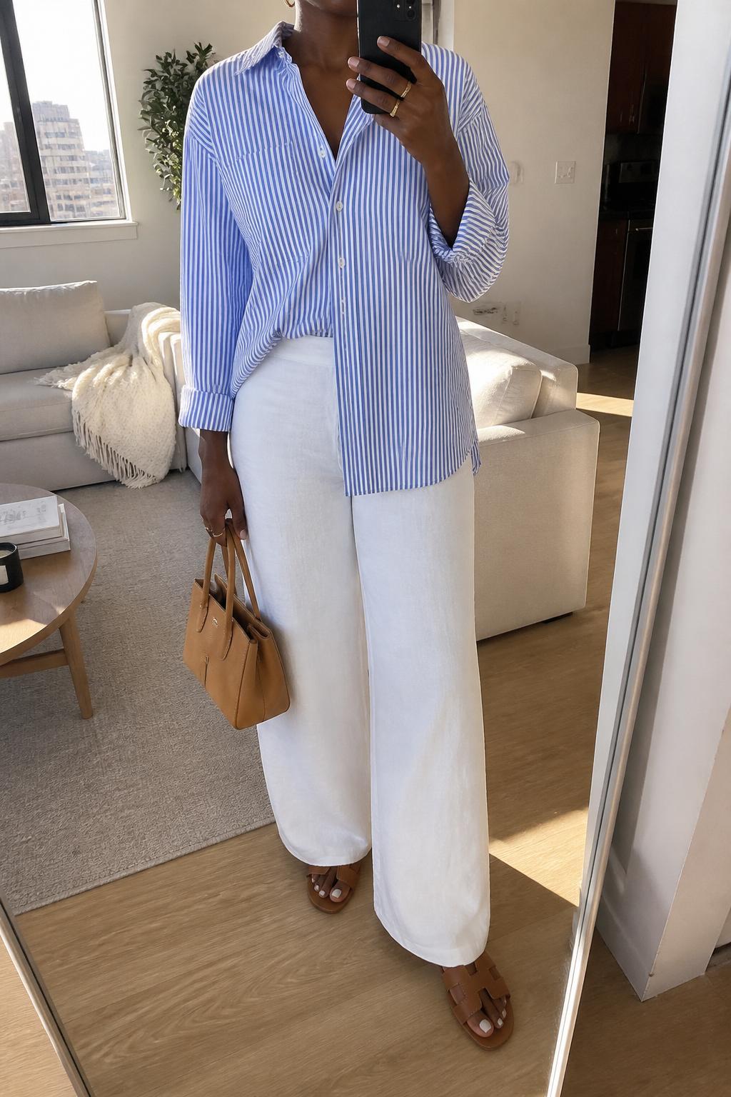 IMAGE_PROMPT: mirror selfie of a black woman wearing a blue and white striped long-sleeve cotton button-down shirt, white wide-leg linen pants, brown leather flat sandals, tan mini tote bag, and small gold hoop earrings, clearly showing the same clothing items listed in the Outfit Pieces section, holding a phone in front of a mirror, cropped at the NECK so the face does not show, relaxed casual selfie pose, in a stylish city apartment living room, soft morning light, with throw blanket on a living room sofa, latest iPhone photo quality.