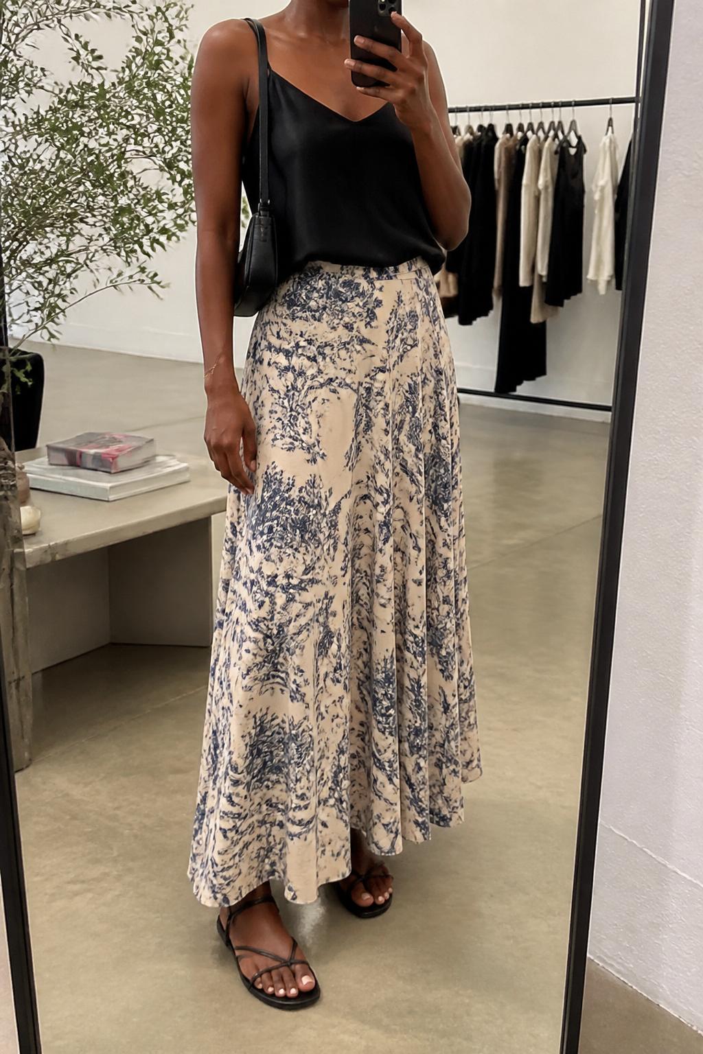 IMAGE_PROMPT: mirror selfie of a black woman wearing a black satin camisole, cream and blue printed flowy maxi skirt, black strappy flat sandals, small black shoulder bag, and delicate gold bracelet, clearly showing the same clothing items listed in the Outfit Pieces section, holding a phone in front of a mirror, cropped at the NECK so the face does not show, relaxed casual selfie pose, in a fashion studio mirror corner, soft indoor lighting, with clothing rack with garments behind, latest iPhone photo quality.