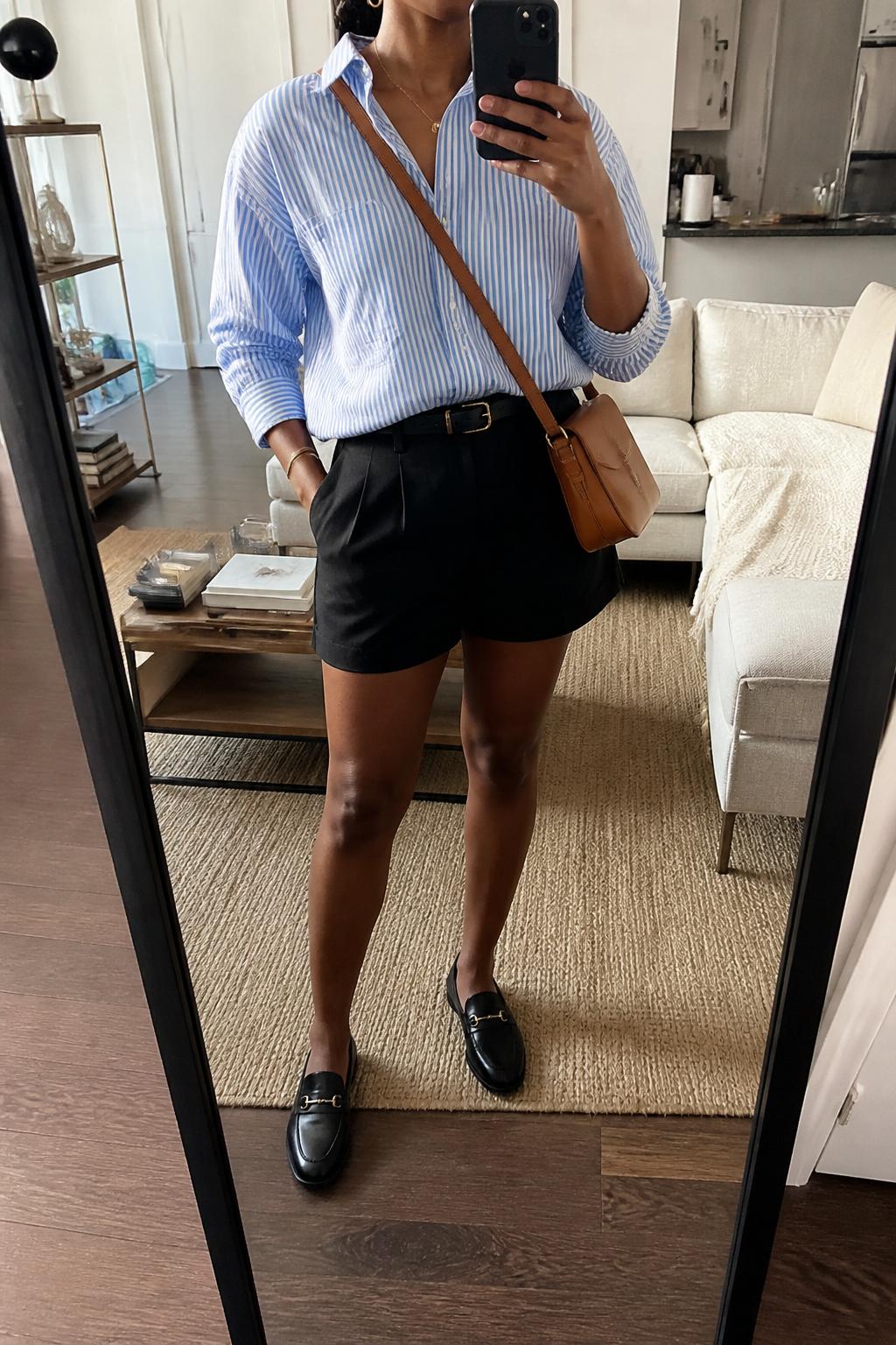 IMAGE_PROMPT: mirror selfie of a black woman wearing a blue and white striped cotton button-down shirt, black tailored shorts, black leather loafers, tan crossbody bag, and small gold hoop earrings, clearly showing the same clothing items listed in the Outfit Pieces section, holding a phone in front of a mirror, cropped at the NECK so the face does not show, relaxed casual selfie pose, in a stylish city apartment living room, soft morning light, with throw blanket on a living room sofa, latest iPhone photo quality.