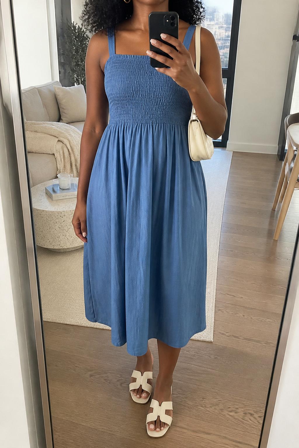 IMAGE_PROMPT: mirror selfie of a black woman wearing a blue smocked cotton sundress, white flat sandals, cream mini shoulder bag, small gold hoop earrings, and soft natural curls, clearly showing the same clothing items listed in the Outfit Pieces section, holding a phone in front of a mirror, cropped at the NECK so the face does not show, relaxed casual selfie pose, in a stylish city apartment living room, soft morning light, with throw blanket on a living room sofa, latest iPhone photo quality.