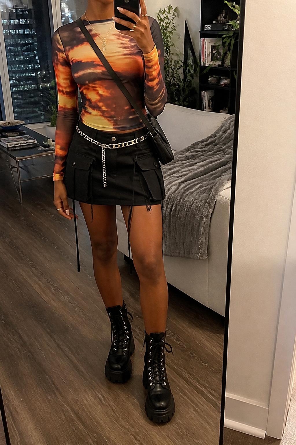 IMAGE_PROMPT: mirror selfie of a black woman wearing a sunset print mesh long-sleeve top, black cargo mini skirt, black chunky combat boots, silver chain belt, and black nylon crossbody bag, clearly showing the same clothing items listed in the Outfit Pieces section, holding a phone in front of a mirror, cropped at the NECK so the face does not show, relaxed casual selfie pose, in a stylish city apartment living room, soft indoor lighting, with throw blanket on a living room sofa, latest iPhone photo quality.