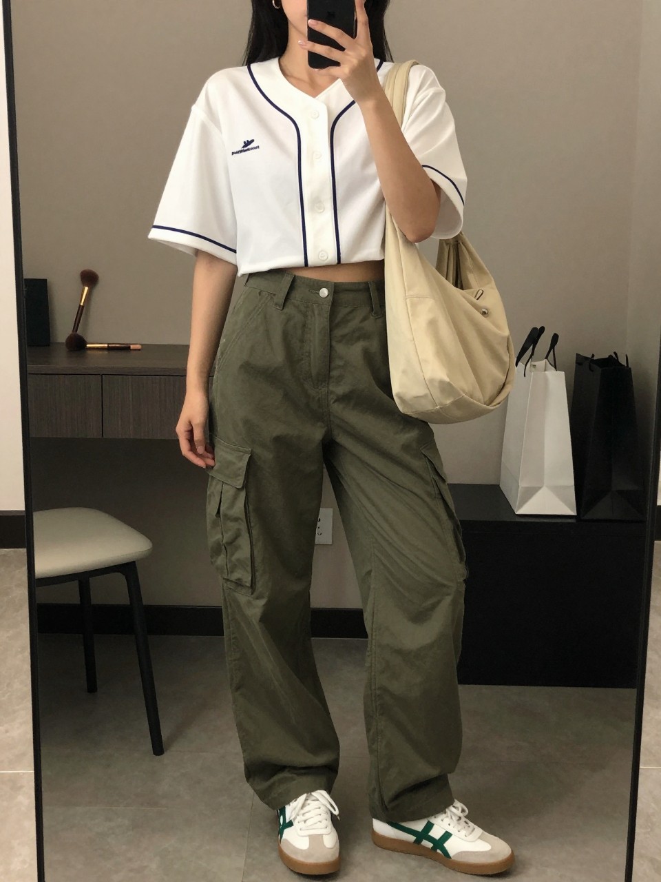 mirror selfie of a woman wearing a cropped white button-front baseball jersey, olive green relaxed cargo pants, white and green retro sneakers, beige nylon shoulder bag, and small gold hoop earrings, clearly showing the same clothing items, colors, fabrics, and accessories listed in the Outfit Pieces section, holding a phone in front of a mirror, cropped at NECK so the face does not show, with a relaxed casual selfie pose, in a fashion studio mirror corner, warm moody lighting, including a makeup brush on the counter and shopping bags beside a chair, latest iPhone photo quality.
