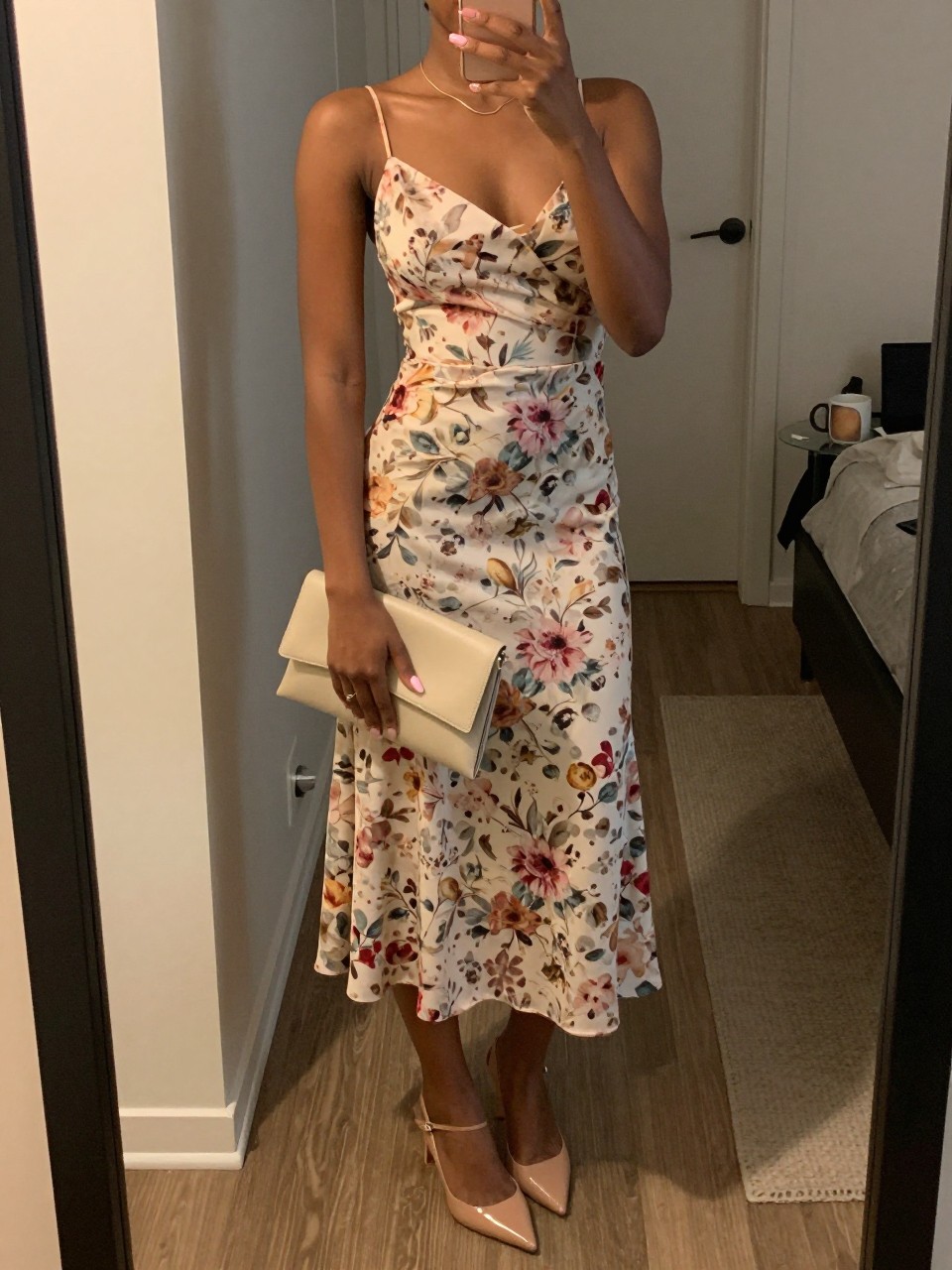 mirror selfie of a black woman wearing a soft floral printed midi dress with nude pointed slingback heels, a thin gold necklace, a cream neutral clutch, and a soft pink manicure, holding a phone in front of a mirror, cropped at NECK so face doesn't show, with a natural casual selfie pose, in a city apartment, warm moody lighting, including small lived-in details like a coffee mug nearby and an open closet behind, latest iPhone photo quality (no watermarks on images)
