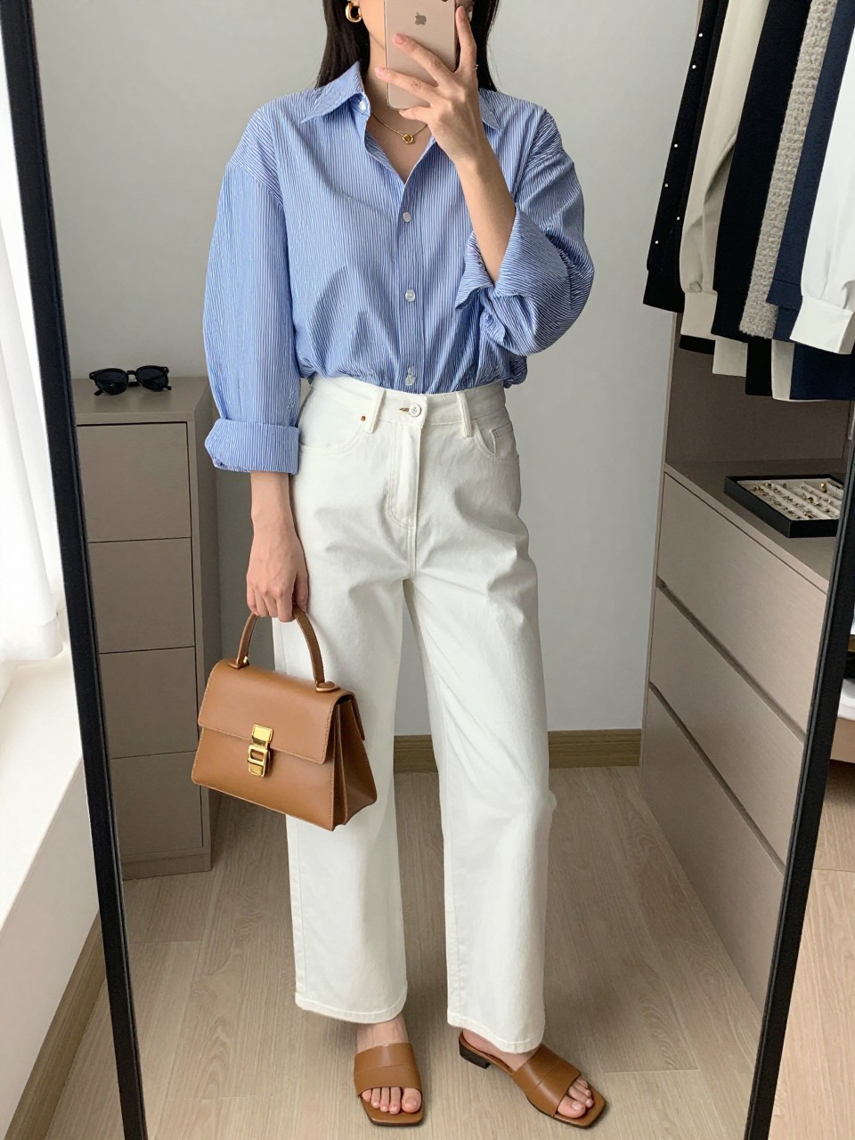 mirror selfie of a woman wearing a blue and white stripe oversized button-down shirt tucked into white high-waisted wide-leg jeans, tan leather slide sandals, tan structured mini bag, and gold hoop earrings, clearly showing the same clothing items, colors, fabrics, and accessories listed in the Outfit Pieces section, holding a phone in front of a mirror, cropped at NECK so the face does not show, with a relaxed casual selfie pose, in a walk-in closet mirror, natural window light, including sunglasses placed on a dresser and a jewelry tray on the vanity, latest iPhone photo quality.