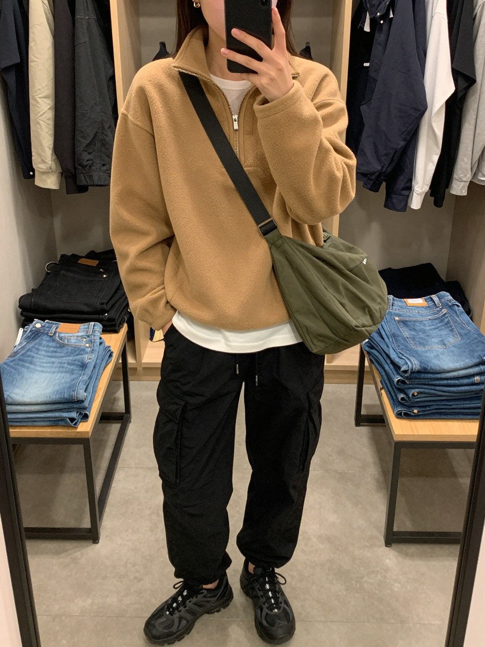 Outfit Image Prompt:
mirror selfie of a woman wearing a camel half-zip fleece pullover, black relaxed cargo pants, white base layer top peeking underneath, black trail sneakers, a dark green nylon crossbody bag, and small gold earrings, holding a phone in front of a mirror, cropped at NECK so the face does not show, relaxed casual selfie pose, in a walk-in closet mirror setup with soft indoor lighting, including a clothing rack with garments behind and folded denim jeans on a bench, natural skin texture, latest iPhone photo quality, realistic and slightly casual.