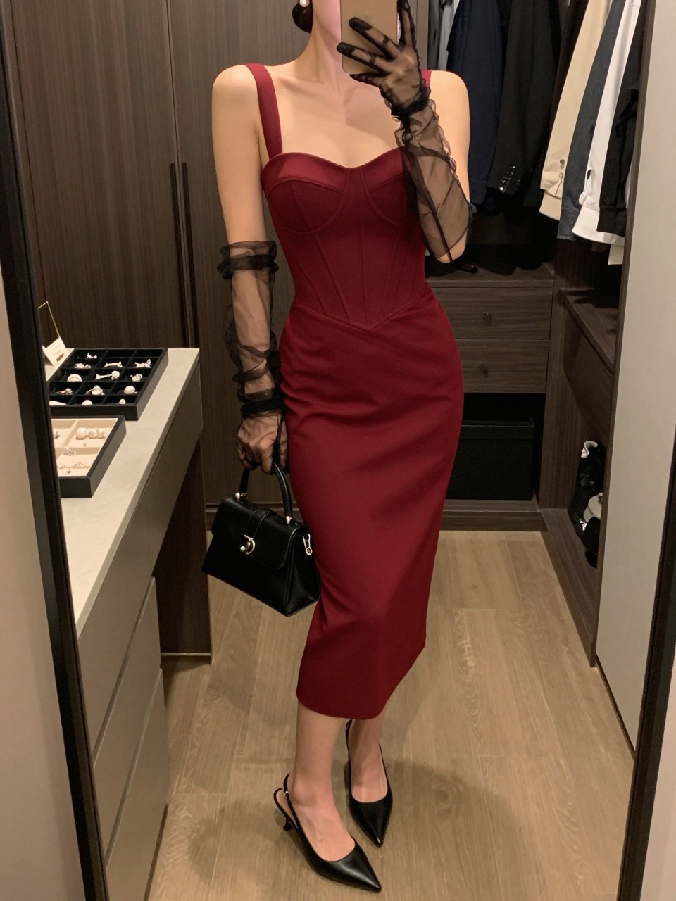 mirror selfie of a woman wearing a garnet red corset midi cocktail dress, sheer black tulle gloves, black pointed-toe slingback heels, pearl stud earrings, a black structured mini bag, and a neat low bun with matte nude makeup, clearly showing the same clothing items, colors, fabrics, and accessories listed in the Outfit Pieces section, holding a phone in front of a mirror, cropped at NECK so the face does not show, with a relaxed casual selfie pose, in a walk-in closet mirror, warm moody lighting, including a jewelry tray on the vanity and open closet with clothes visible, latest iPhone photo quality.